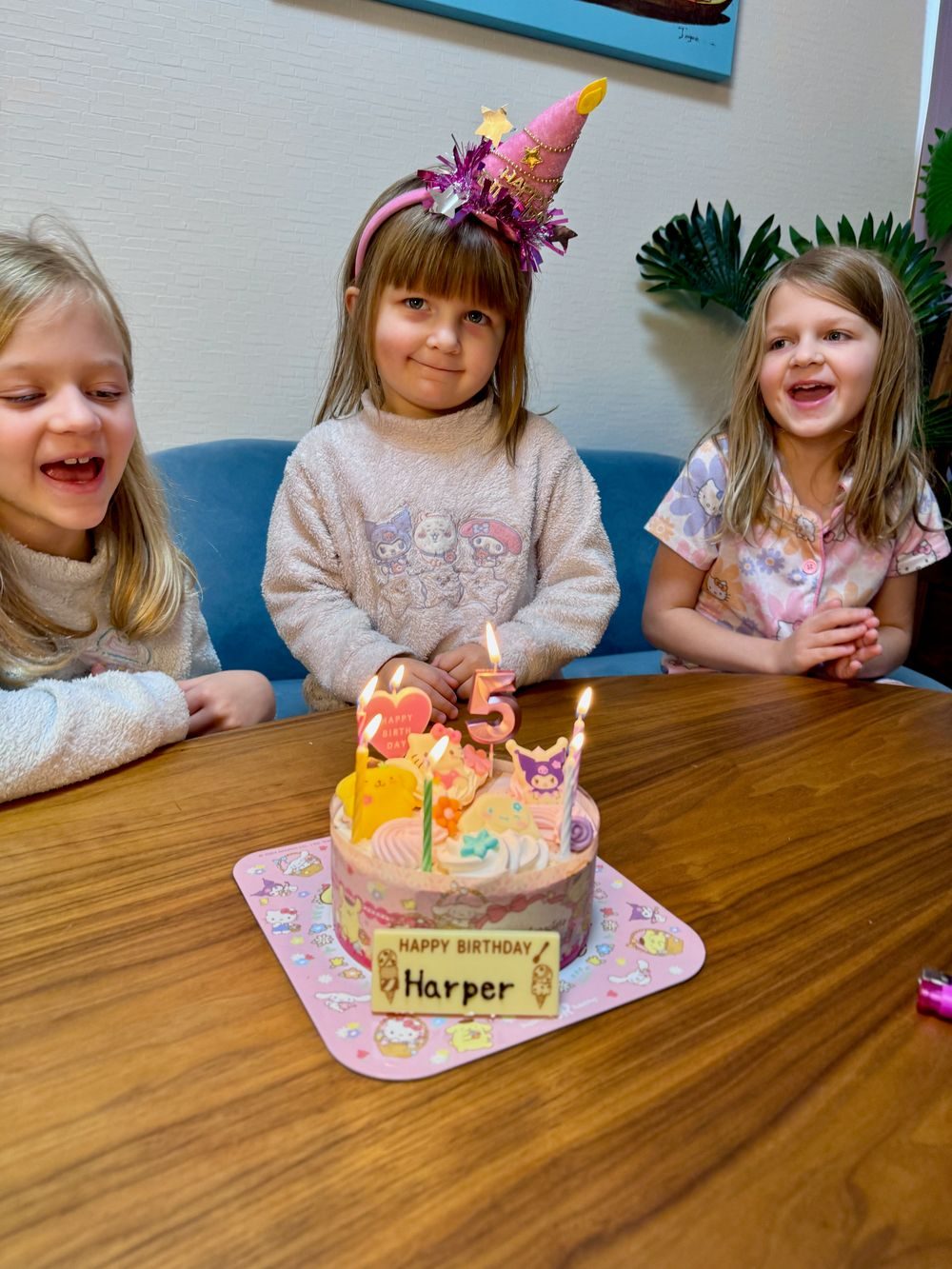 Three young girls, one wearing a birthday hat, sit together in front of a lit birthday cake, two of them laughing.