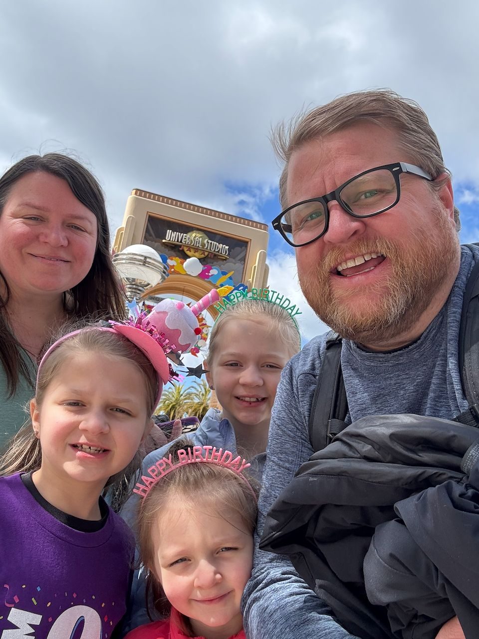 A family selfie in front of the Universal Studios Japan entrance globe, with two girls wearing Happy Birthday headbands, a third girl smiling behind them, and two adults flanking the group under a partly cloudy sky.