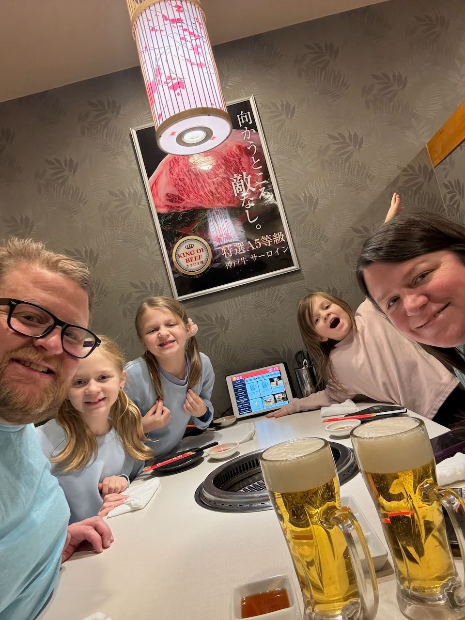 Adam takes a selfie at a yakiniku restaurant table in Osaka with Lindsay and their three daughters, two large beers in the foreground, a tablet ordering system on the table, and an A5 Kobe beef poster on the wall behind them.
