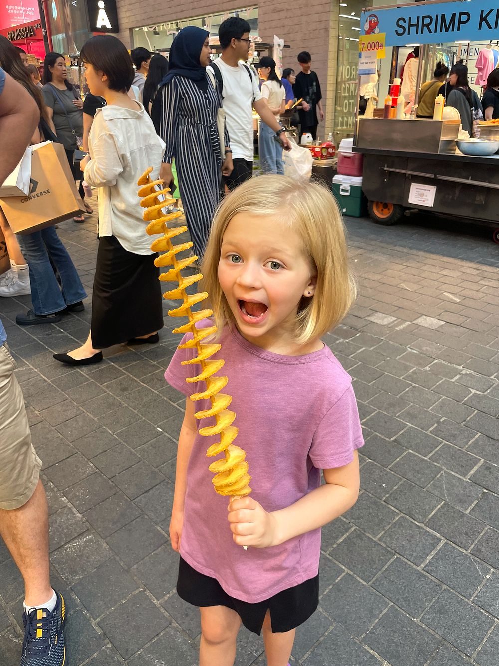 A young girl on a busy Myeongdong street holding a spiral-cut tornado potato on a stick with her mouth open in excitement