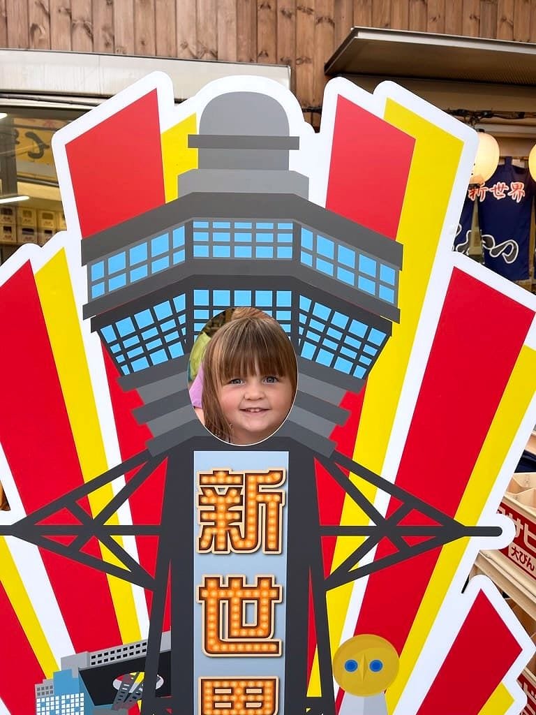 A young girl's face peeking through the hole in a colorful Tsutenkaku tower photo cutout sign in Shinsekai, Osaka, with Japanese shopfronts visible in the background.