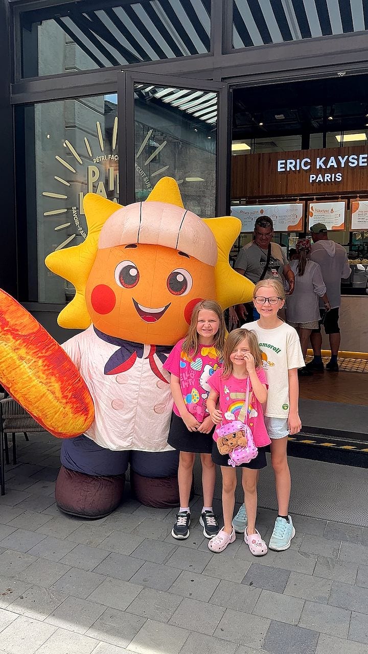 Three girls pose with a large inflatable sun-shaped bakery mascot in front of an Eric Kayser Paris storefront, the youngest rubbing her eye while the other two smile at the camera.