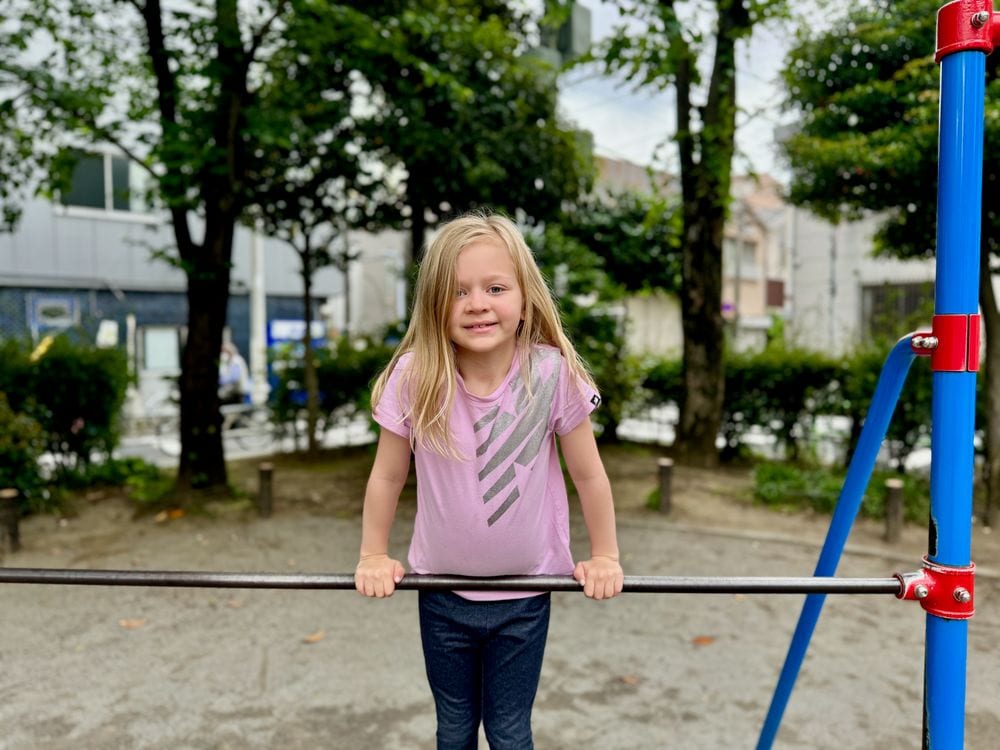 A young girl grips a horizontal bar on playground equipment at Iriya Minami Park, smiling at the camera with trees in the background.
