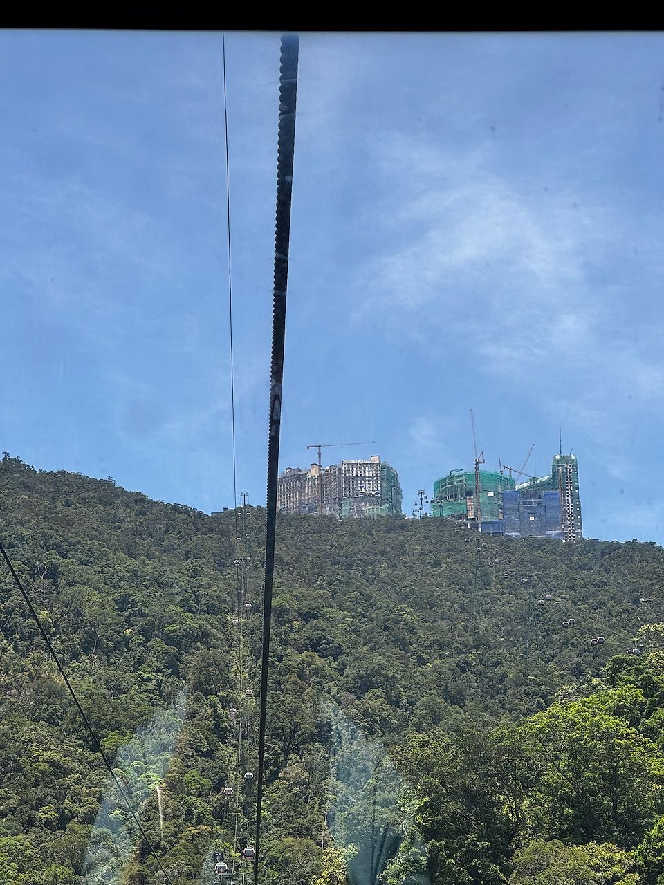 View through a cable car window of a forested hillside at Ba Na Hills with several large hotel buildings under construction and cranes on the summit