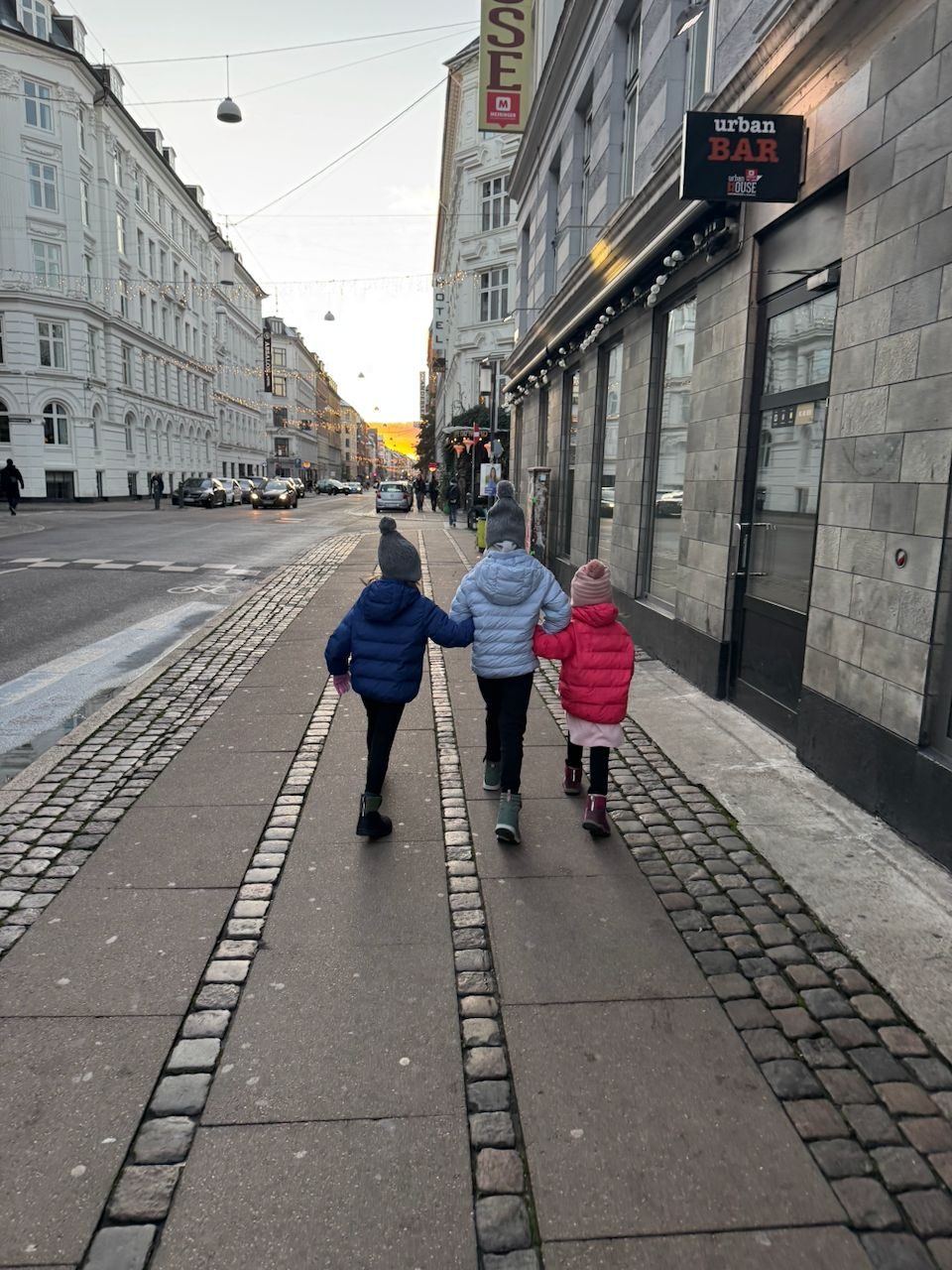 Three girls holding hands walking away from the camera through central Copenhagen at dusk