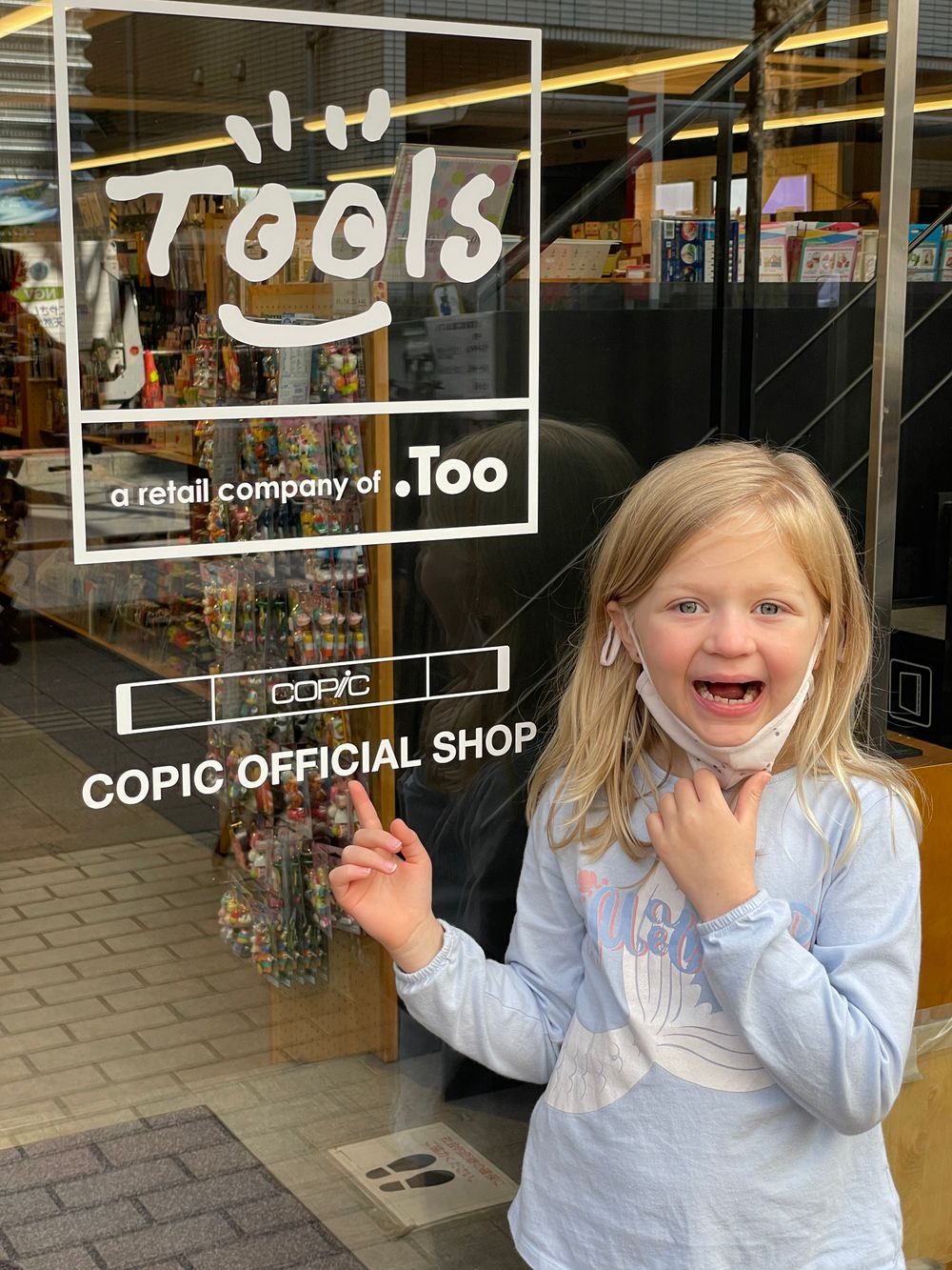 A young girl points excitedly at the glass storefront of the Copic Official Shop in Tokyo, decorated with the TOOLS and COPIC logos.