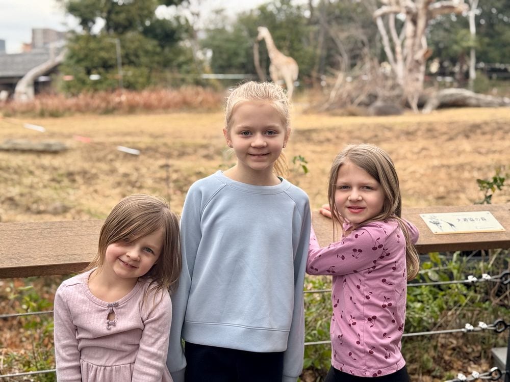 Lily, Harper, and Cora standing at a railing at Tennoji Zoo in Osaka with a giraffe visible in the enclosure behind them
