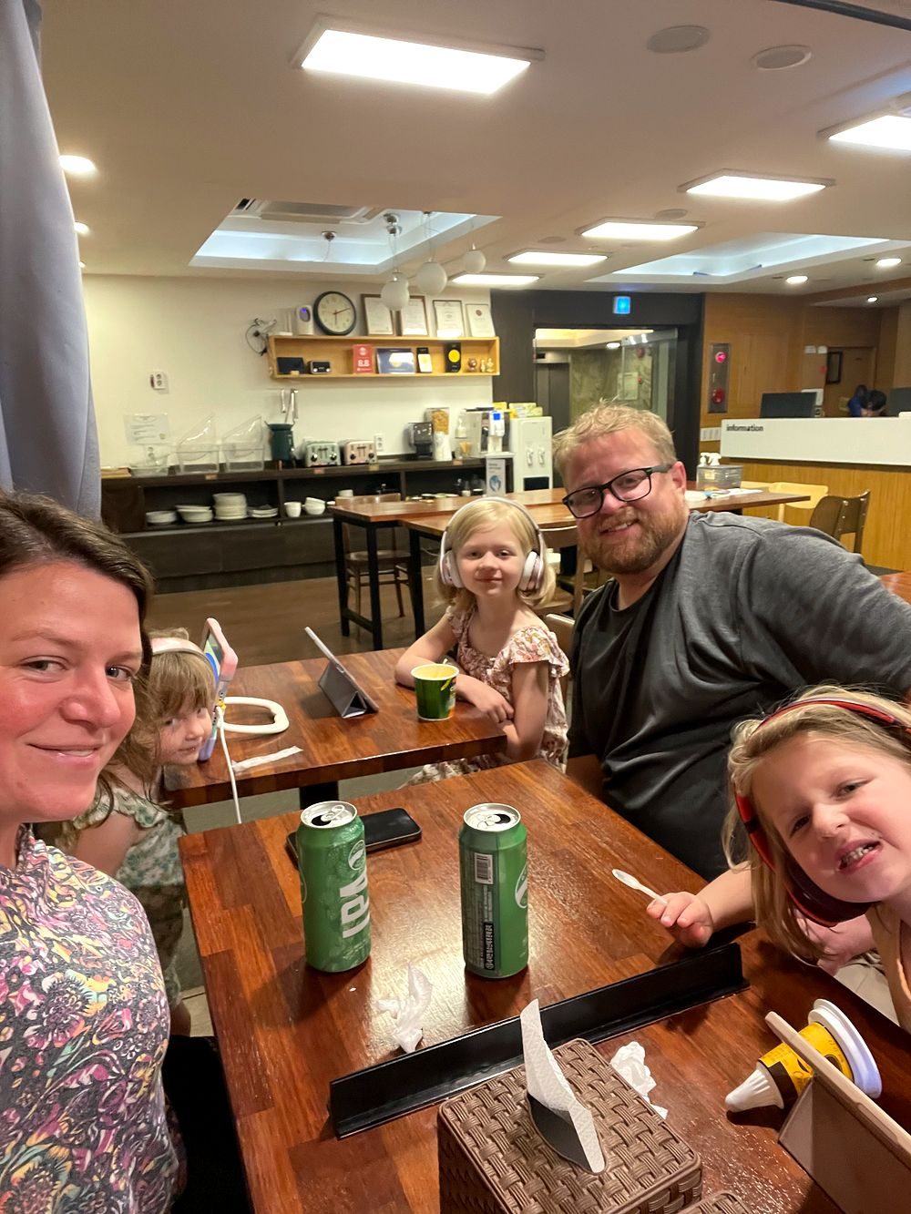 Family selfie around a wooden table in a Seoul hostel common room with two Goose Island IPA cans on the table and kids wearing headphones