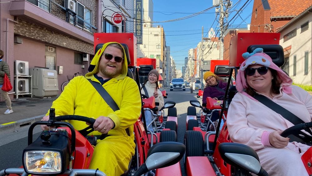 Adam in a yellow Pikachu onesie and Lindsay in a pink My Melody onesie drive open-air go-karts through a narrow street in Nishi-Asakusa, Tokyo, both wearing sunglasses and smiling at the camera with other kart riders visible behind them.