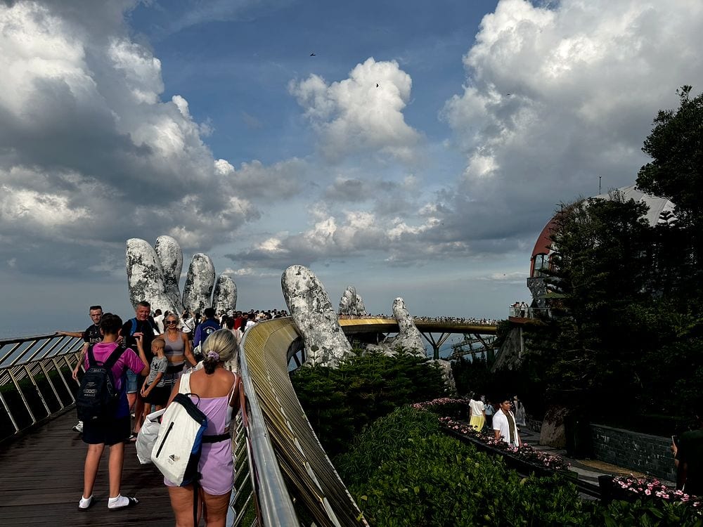 A wide shot looking along the Golden Bridge at Ba Na Hills, with two large weathered stone hands holding the curved wooden walkway above the mountain treeline, crowds of tourists walking in both directions under a dramatic sky of blue and white clouds.