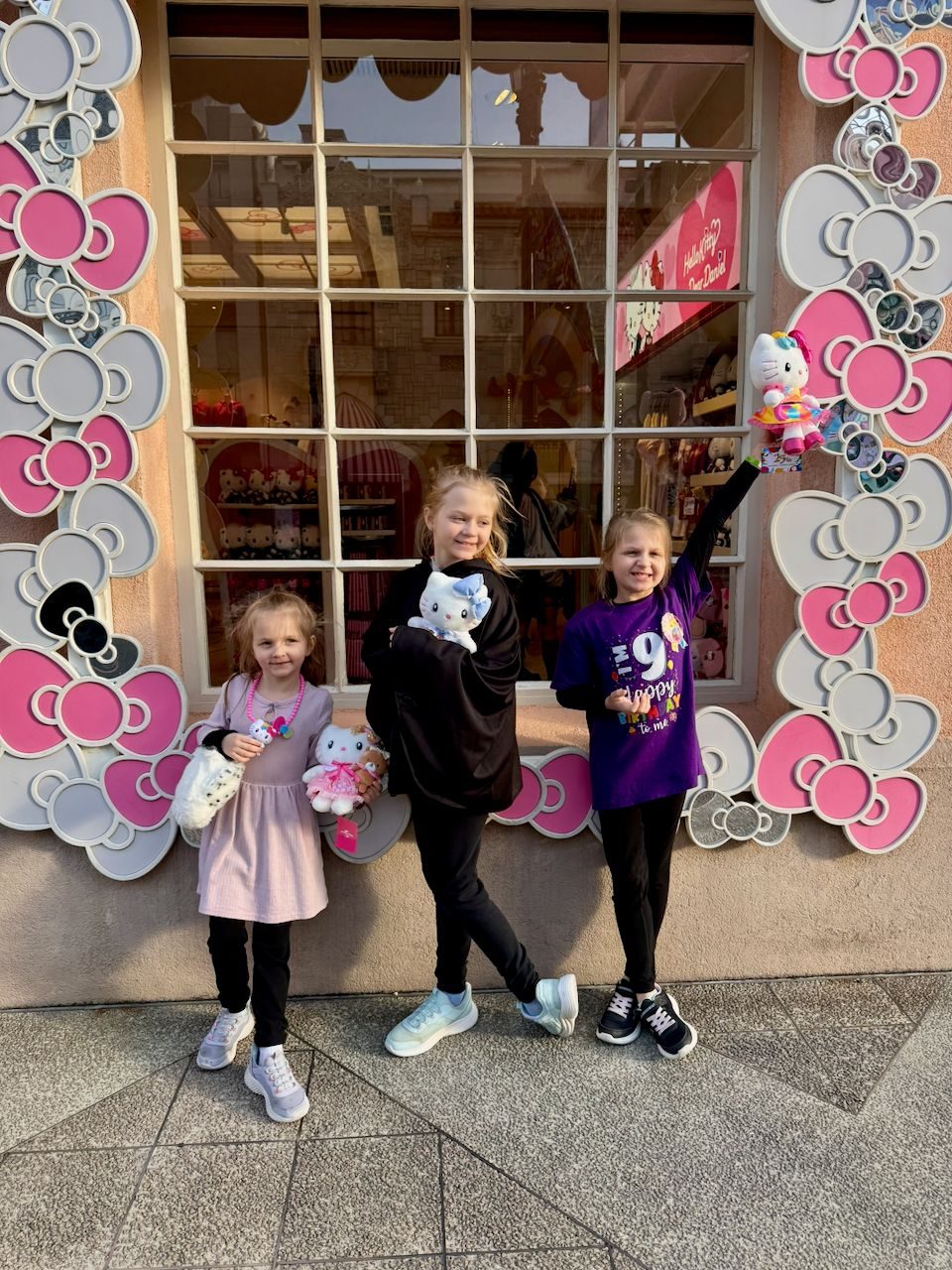 Lily, Cora, and Harper stand in front of a Hello Kitty store at Universal Studios Japan, each holding Hello Kitty plush toys, with Cora wearing a purple Happy 9th Birthday to Me shirt and Harper raising her plush in the air.