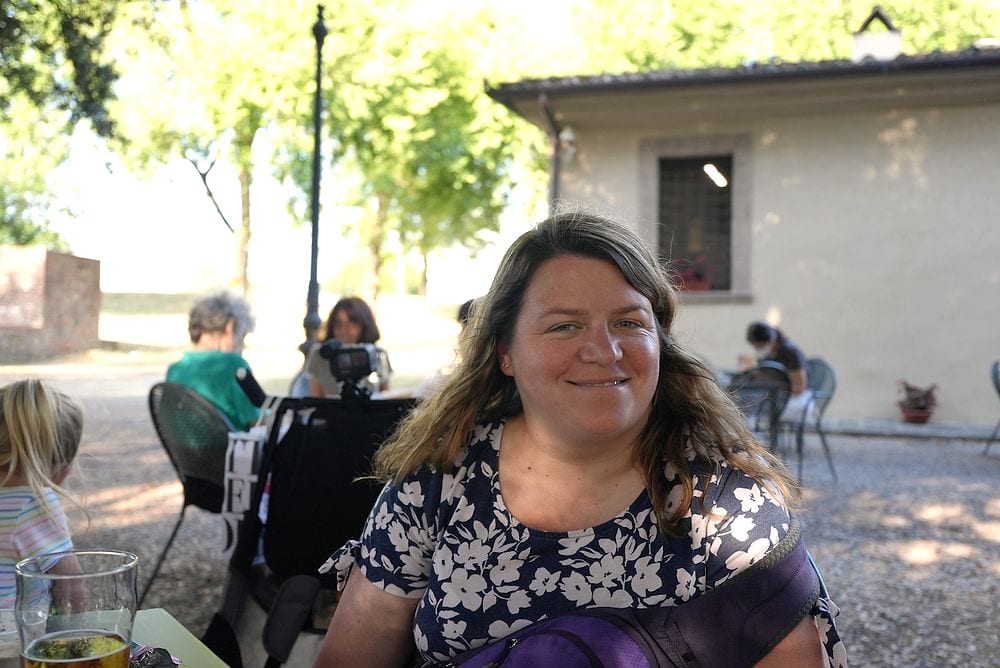 Lindsay sits at an outdoor cafe table on the Lucca city walls, smiling at the camera, with a tree-lined gravel path and stone buildings behind her