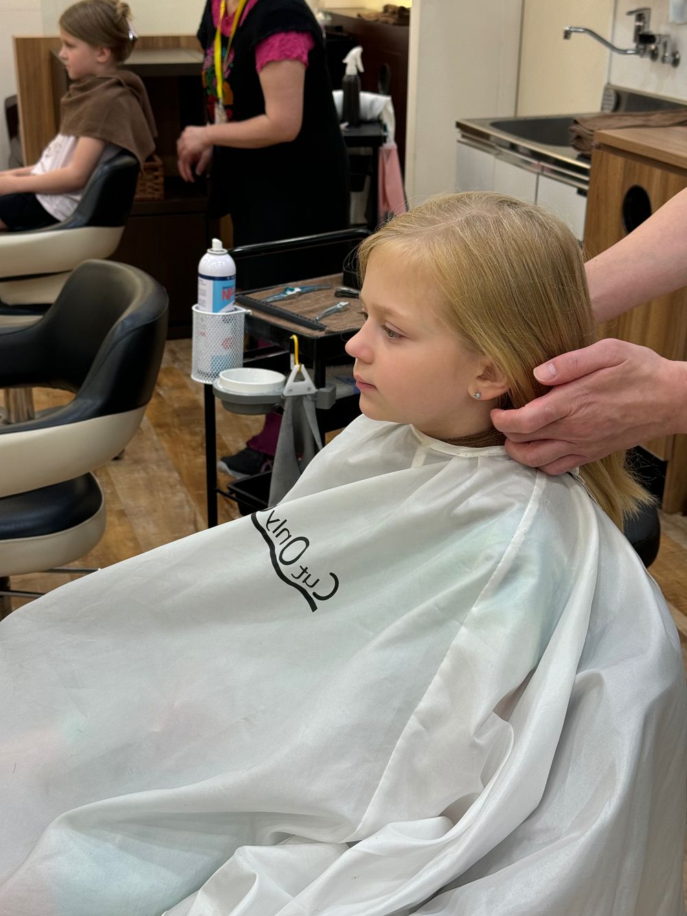 A young girl sits in a salon chair wearing a plain white cutting cape while a stylist's hands are visible working on her hair.