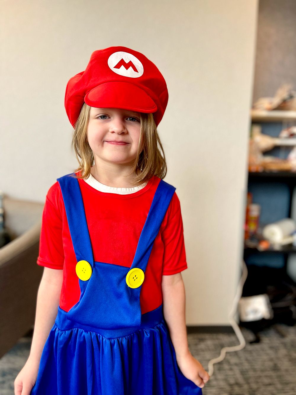 A young girl wears a red Mario costume with a red cap featuring the M logo, red shirt, and blue overalls.