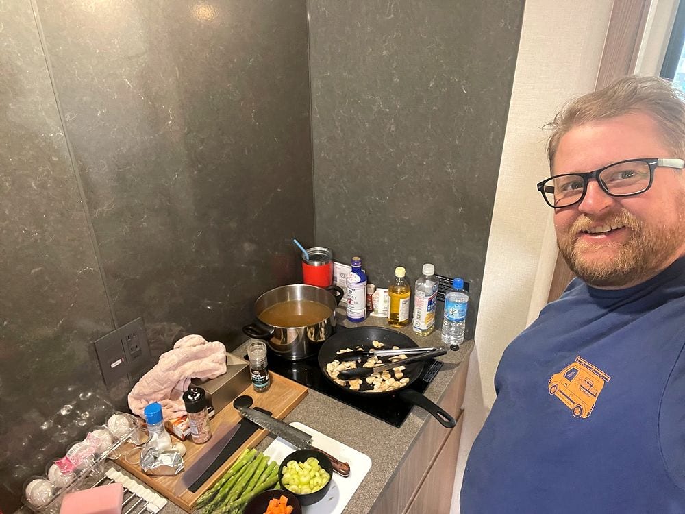A man smiles while cooking at a small kitchen counter in a Tokyo apartment, with pans, vegetables, and seasonings laid out in front of him.