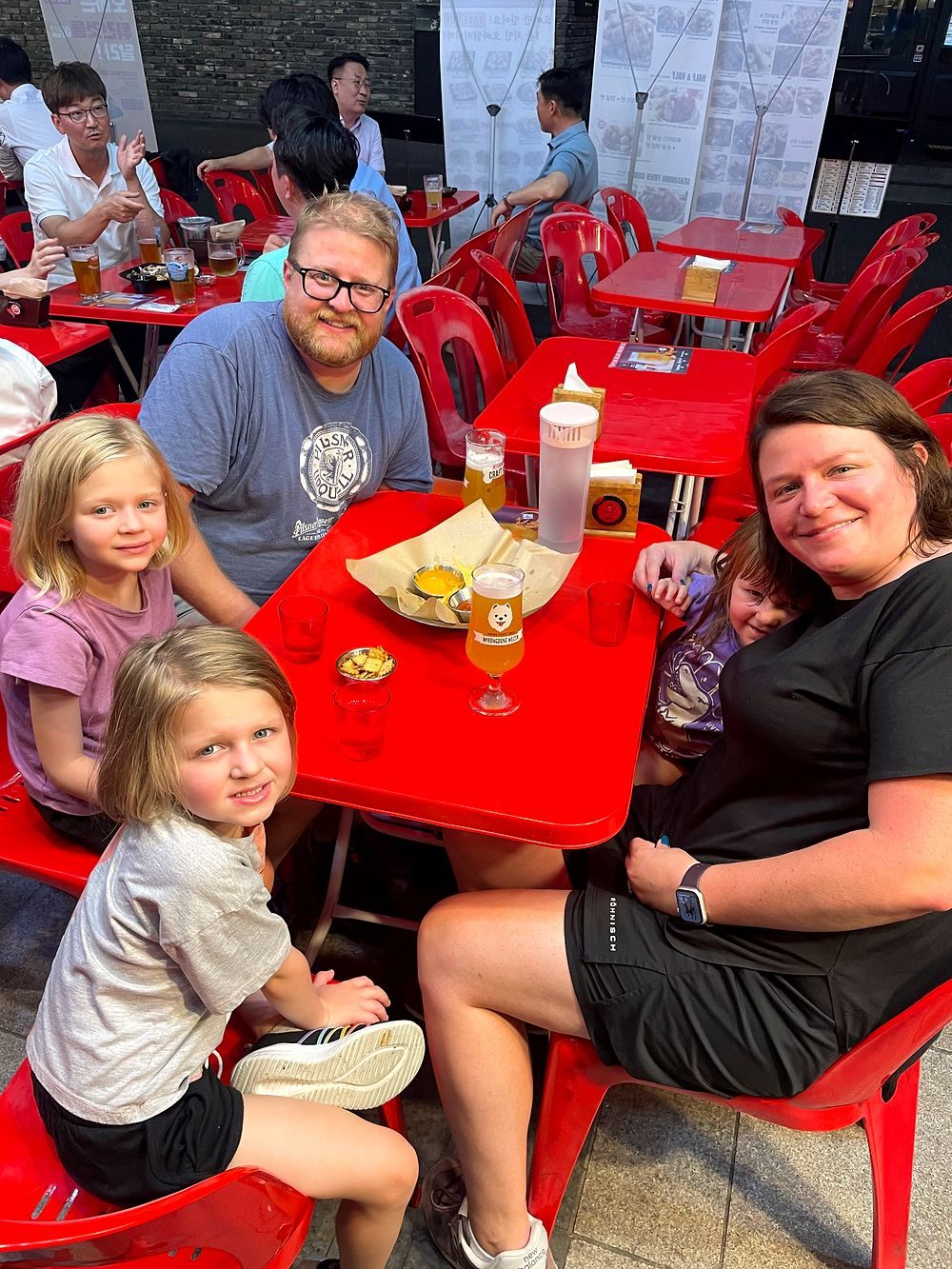 A family of five at a red plastic table at an outdoor Seoul beer hall in the evening with a draft beer and snacks