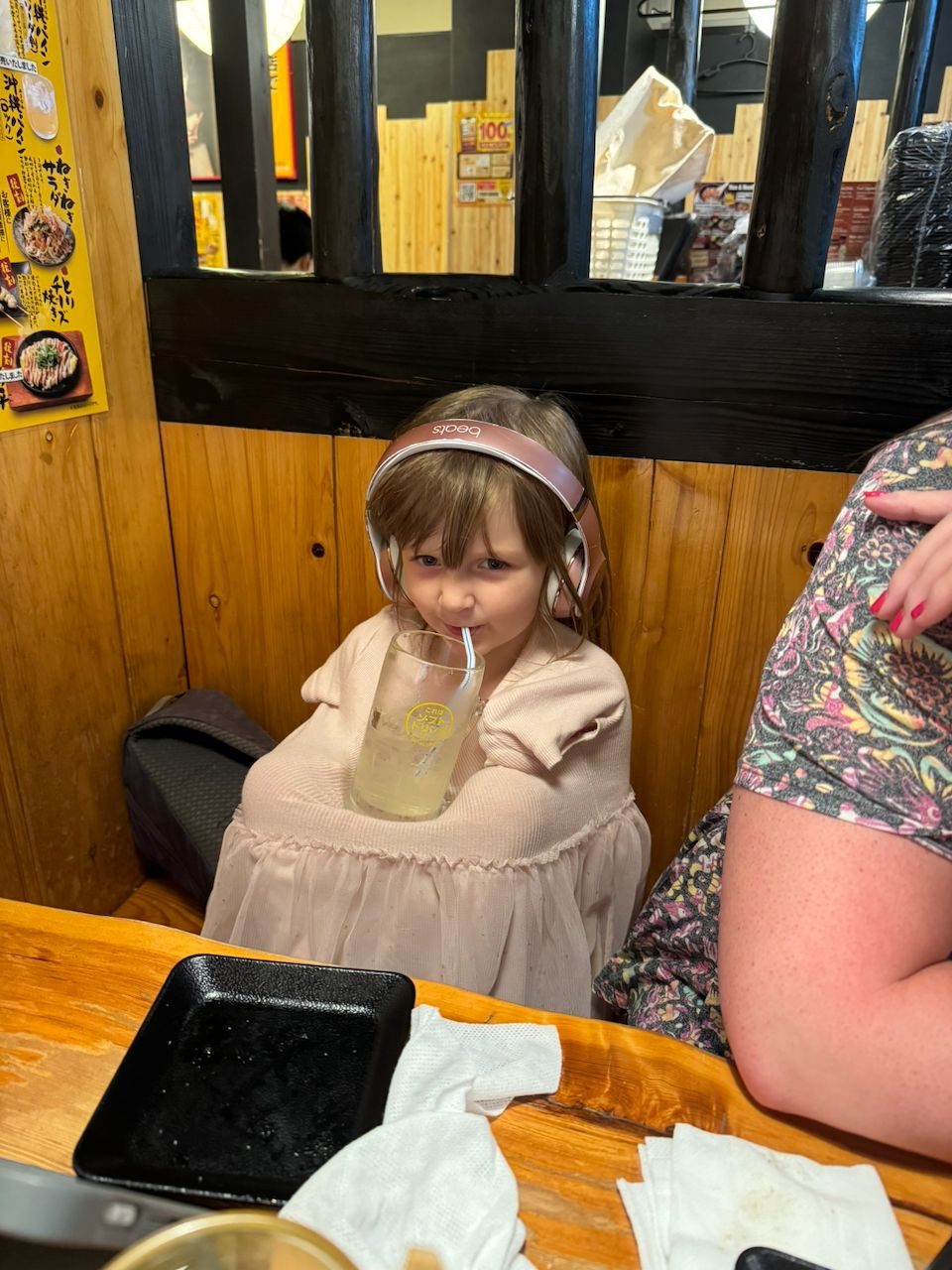 A young girl wearing pink Beats headphones sips a lemonade through a straw at a wooden booth inside a Japanese izakaya, looking directly at the camera with a relaxed expression.