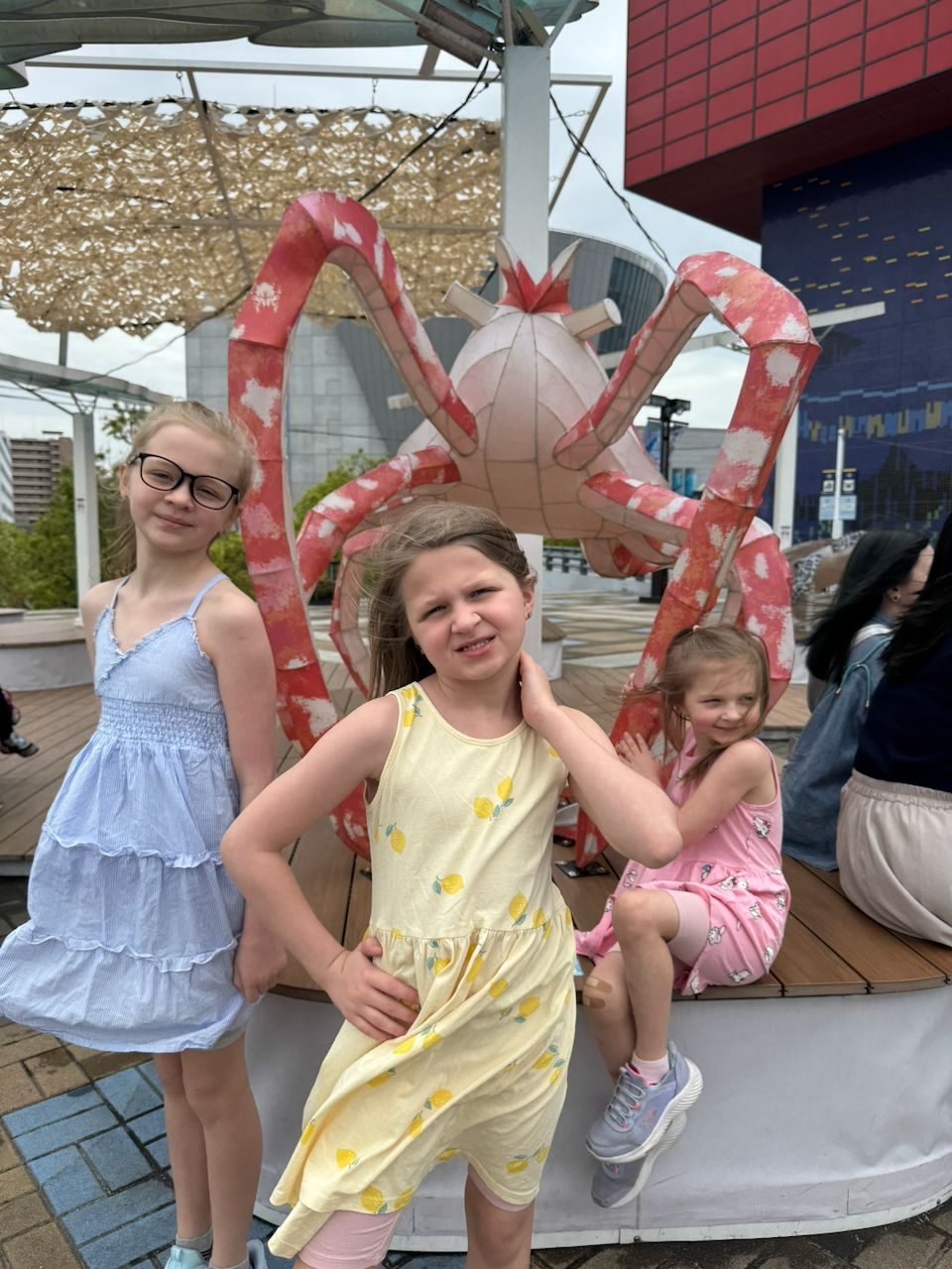 Three girls pose in front of a large decorative spider crab sculpture on an outdoor waterfront plaza in Osaka.