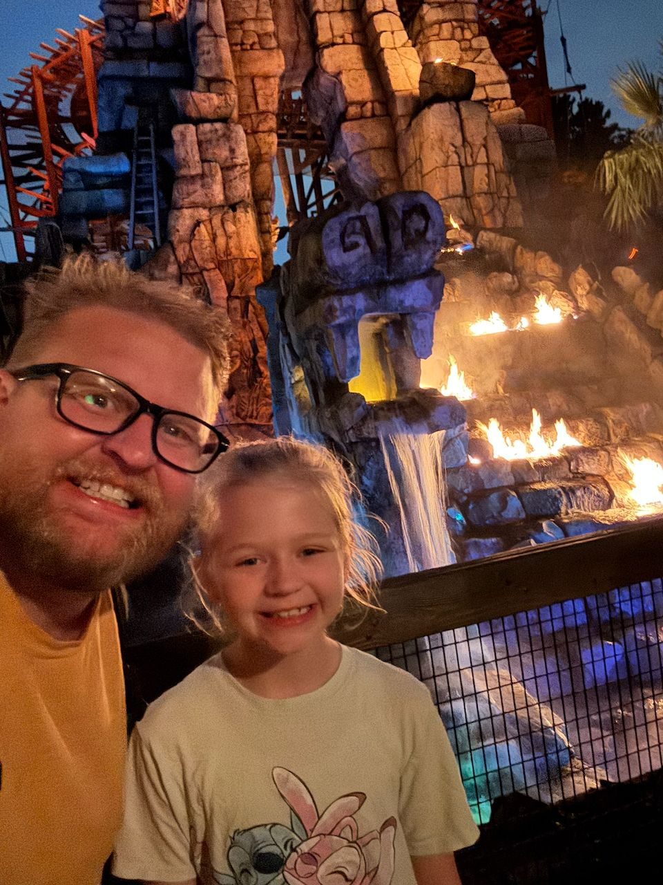 A man and young girl smile at the camera in front of a towering fake volcano with real fire and waterfall effects at Tokyo DisneySea's Mysterious Island.