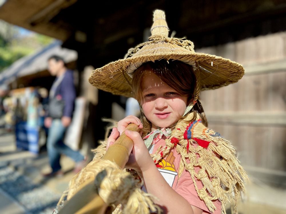 A young girl wearing a large woven straw hat and a traditional Japanese straw cape holds a wooden tool and looks toward the camera, with a thatched historic building blurred in the background.