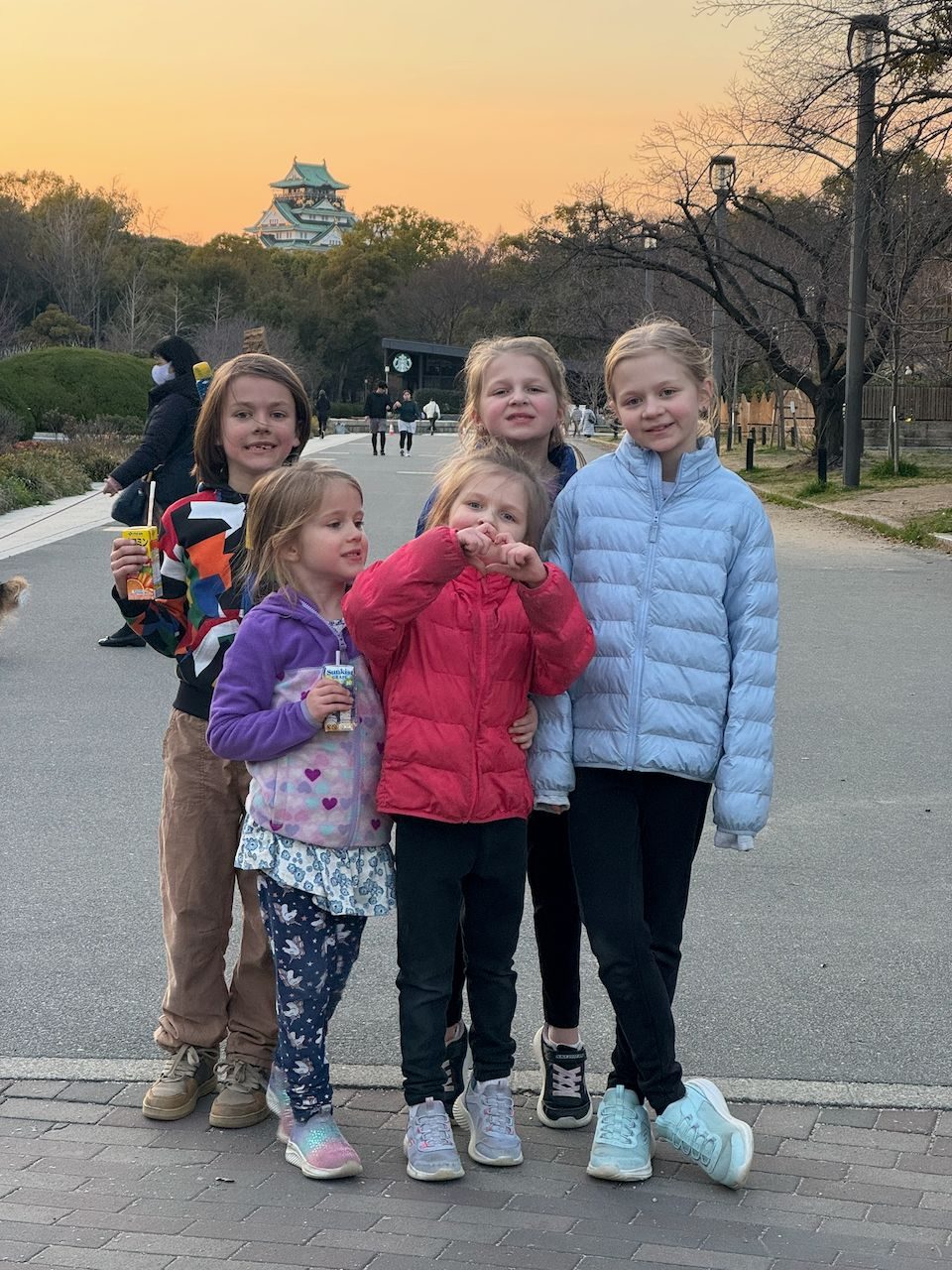 Five girls posing together on a path in Osaka Castle park at sunset with the castle in the warm orange sky behind them