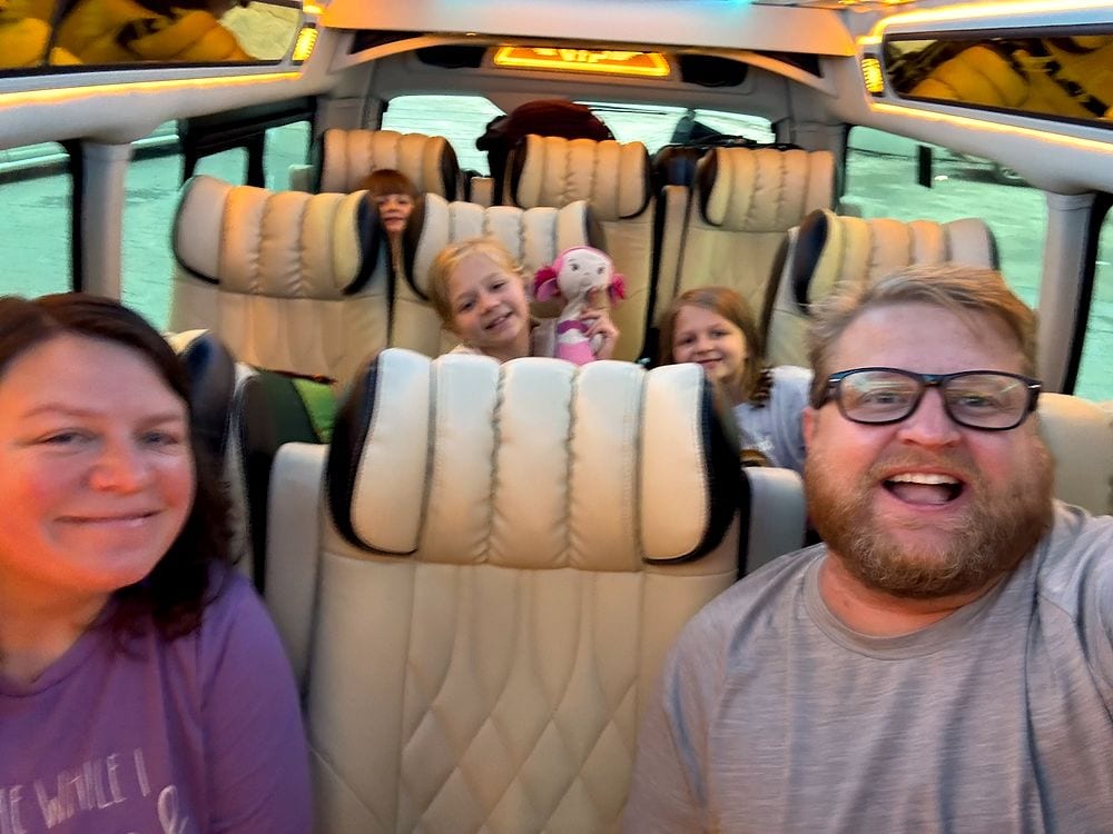 Family selfie of two adults and three girls in a van with cream quilted seats during a daytime transfer in Bangkok, one girl holding a pink stuffed animal