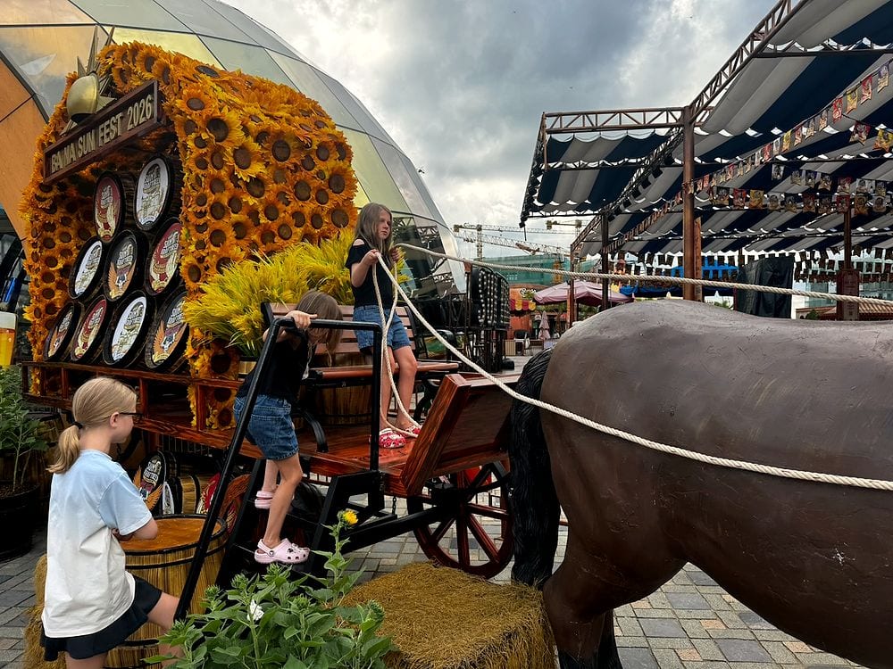 Three girls climb a decorated wooden wagon covered in sunflowers and beer barrels at the Ba Na Sun Fest 2026 installation, with a large dark horse sculpture in the foreground and a geodesic dome structure behind them under a cloudy sky.