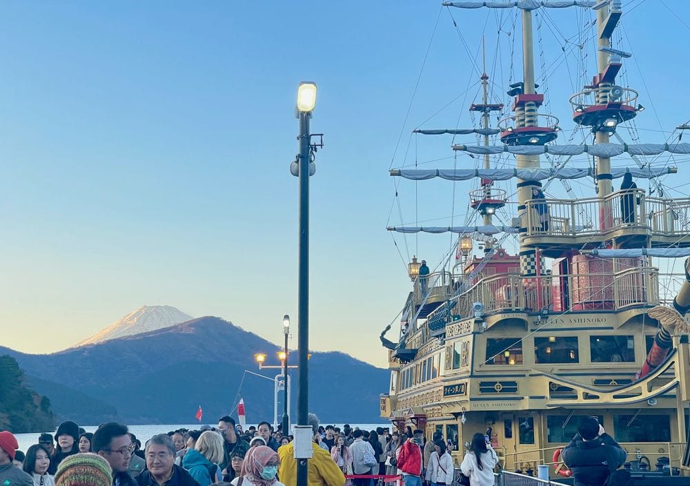 A large ornate pirate-style sightseeing vessel called Queen Ashinoko docked at a crowded pier on Lake Ashi, with Mount Fuji visible in the distance under a clear blue sky at dusk.