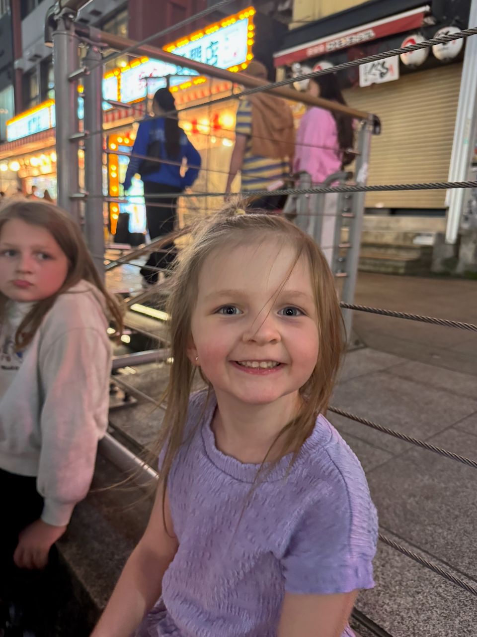 A young girl in a lavender top smiles broadly at the camera from a bridge railing in Dotonbori at night, with neon signs glowing in the background and another child visible to her left.
