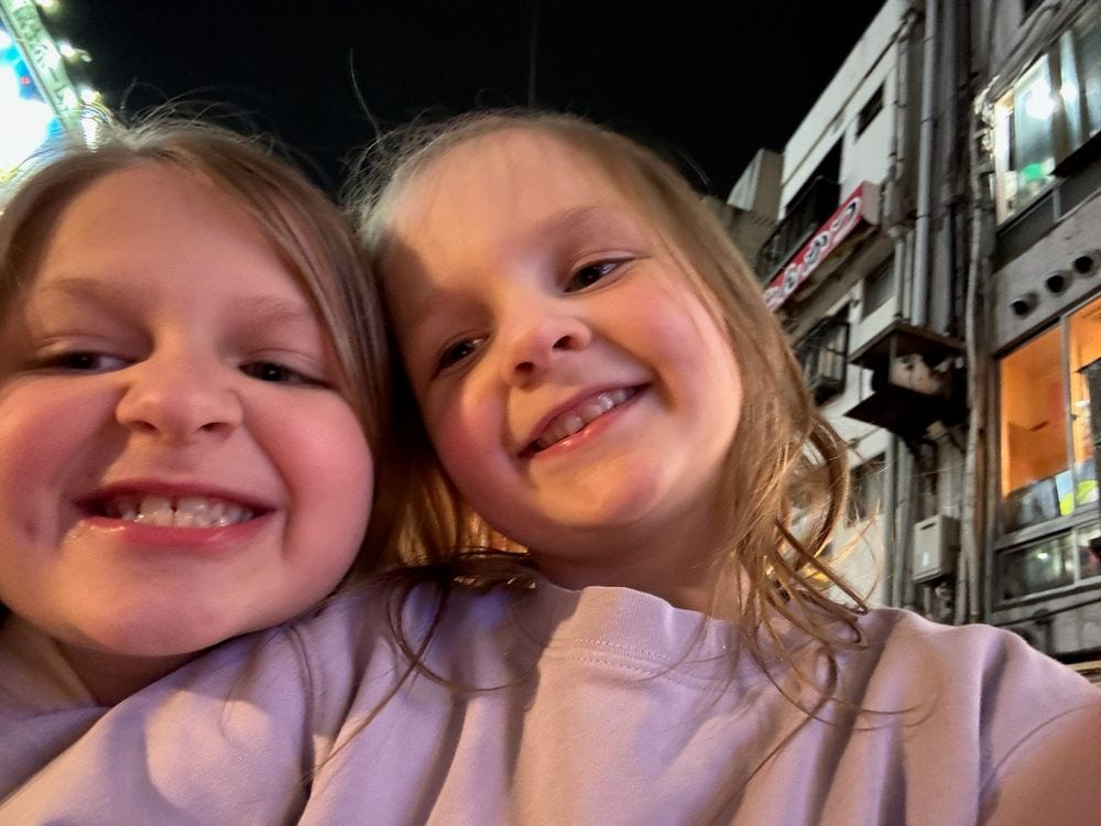Two young girls grinning cheek to cheek at night on a busy Osaka street, with illuminated building signs visible behind them.