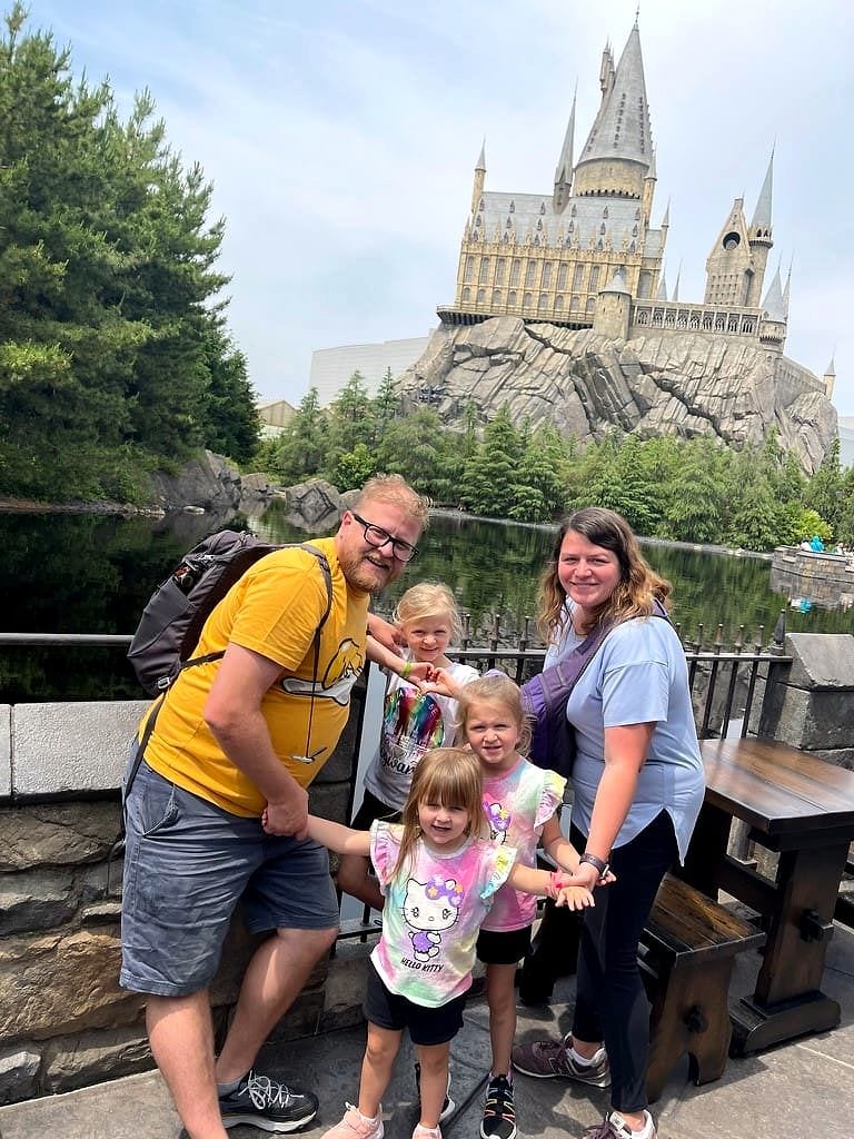 A family of five poses together in front of Hogwarts Castle at the Wizarding World of Harry Potter in Universal Studios Japan, with a reflective pond and pine trees behind them on an overcast day.