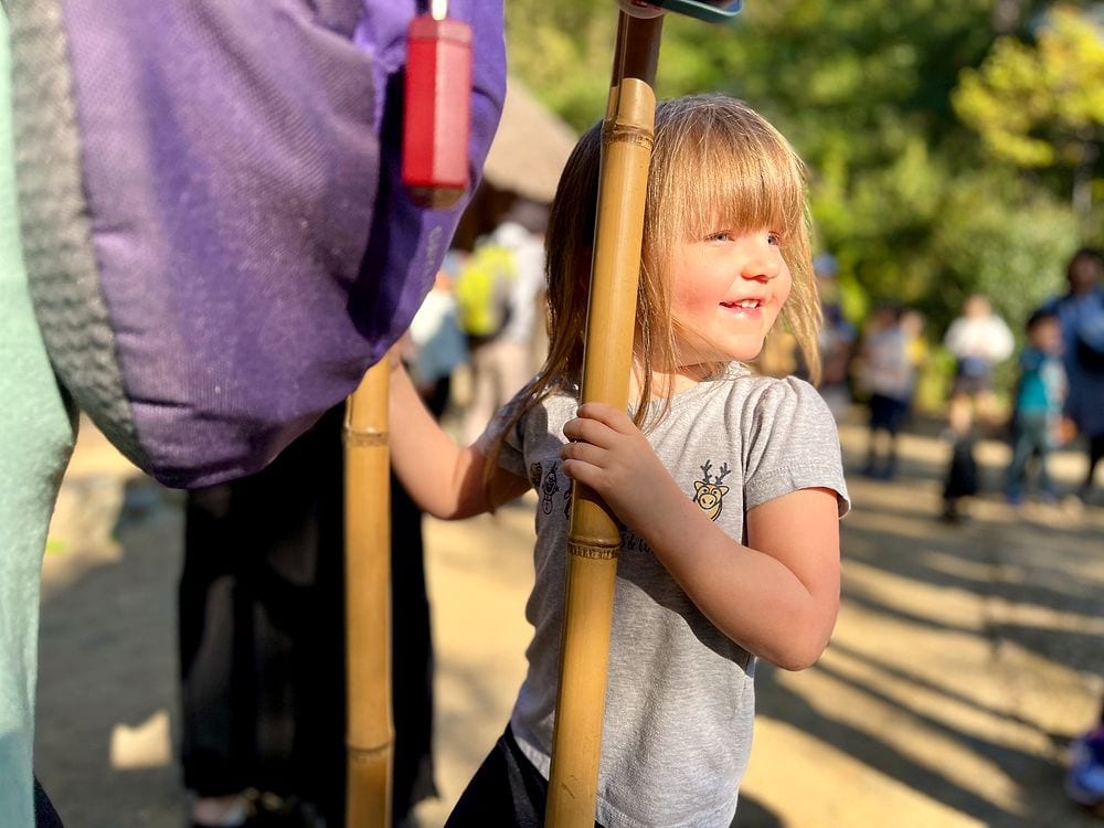 A young girl holds a long bamboo pole alongside an adult at an outdoor cultural site, autumn trees visible behind them.