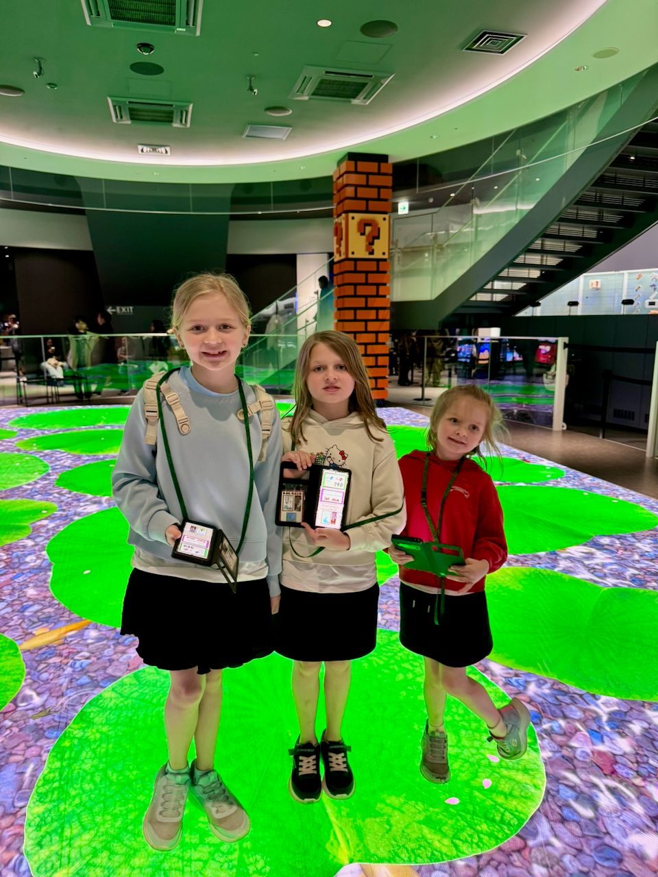 Three girls holding their entry cards at the Nintendo Museum in Uji, standing on a giant interactive Yoshi's Island floor display