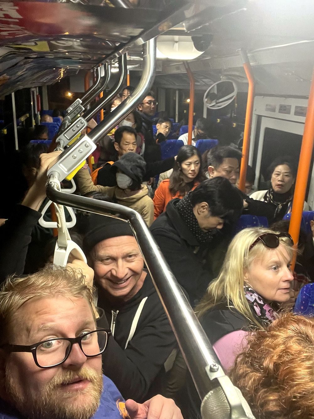 A crowded public bus at night with passengers standing and holding overhead rails, including a man and an older couple visible in the foreground.