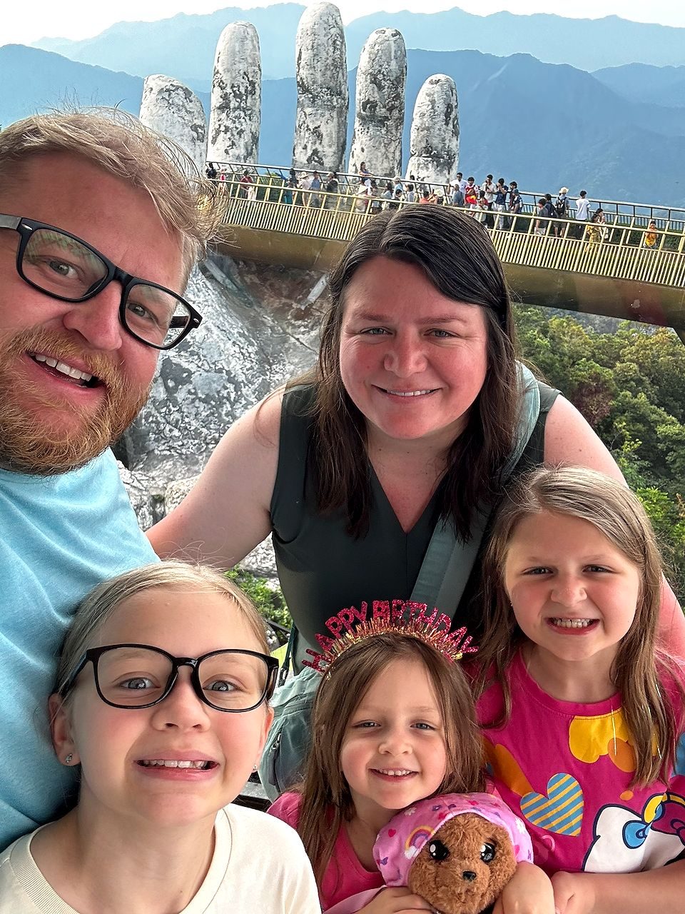 A family of five taking a selfie on the Golden Bridge at Ba Na Hills, with the two giant stone hands holding the bridge behind them, one child wearing a gold birthday crown