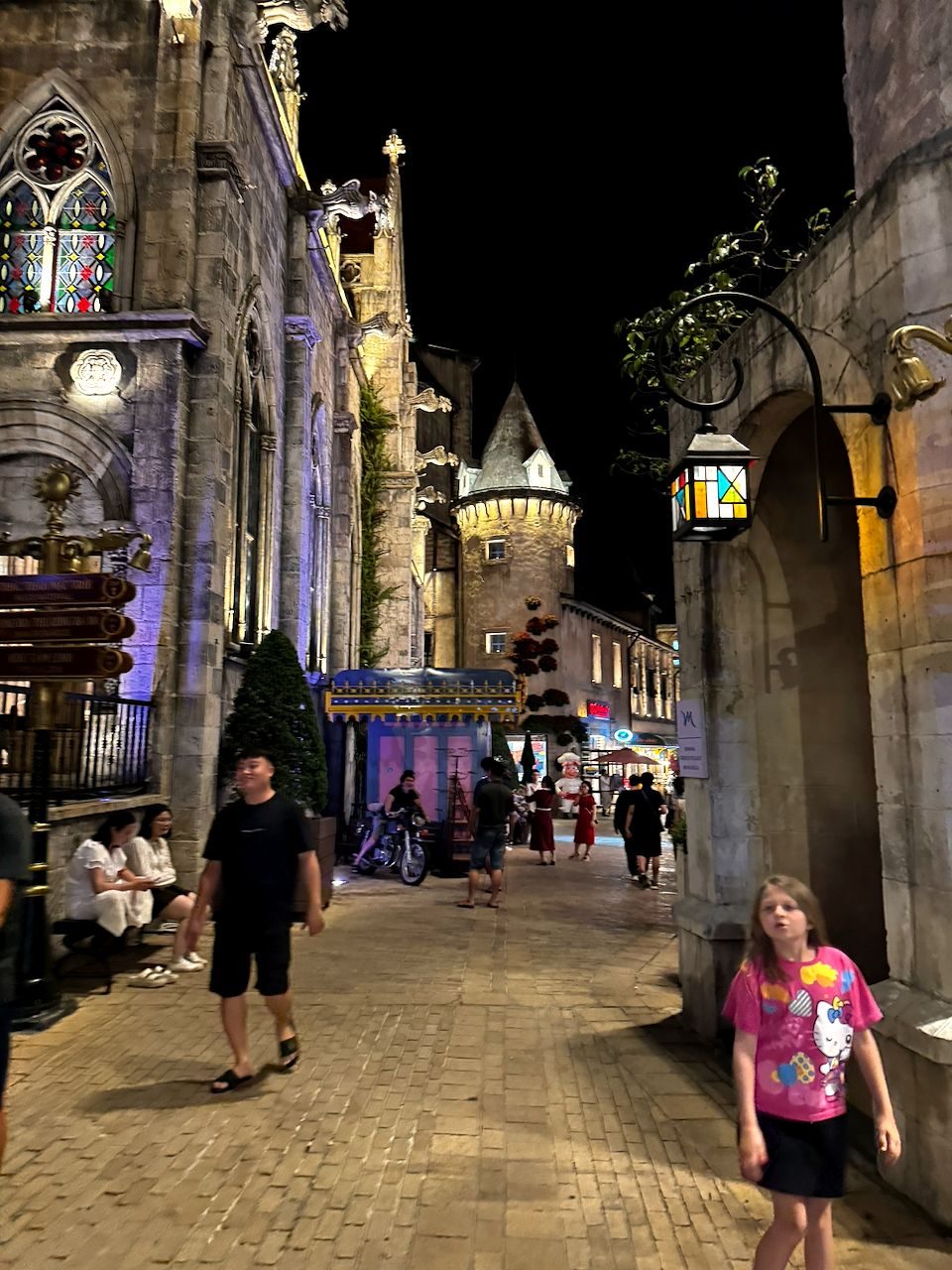 A young girl in a pink Hello Kitty shirt walks at night through a cobblestone alley flanked by illuminated Gothic-style stone buildings at Ba Na Hills in Da Nang, Vietnam.