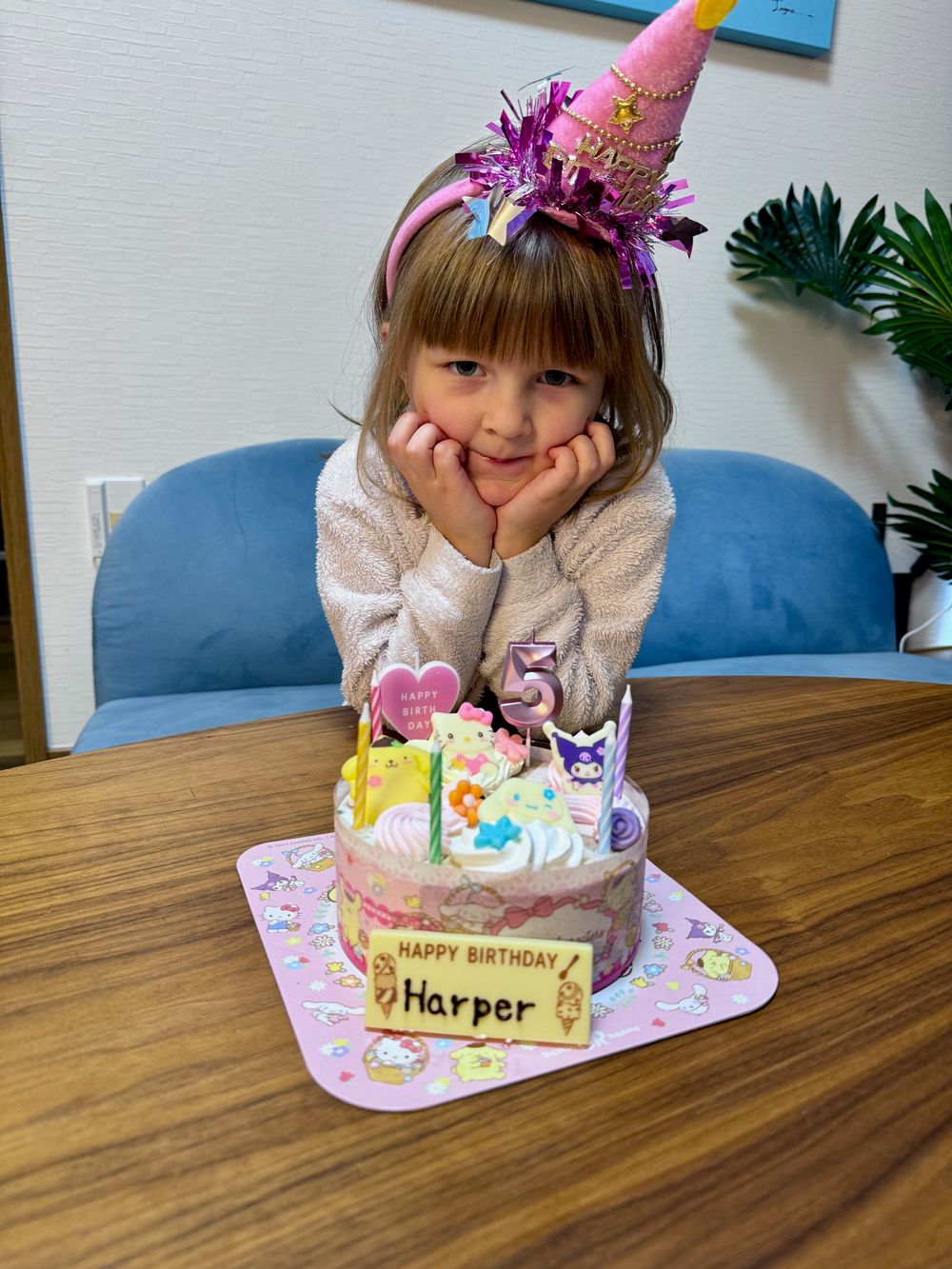 A young girl wearing a pink birthday hat smiles in front of a decorated Sanrio-themed birthday cake with a nameplate reading Happy Birthday Harper.