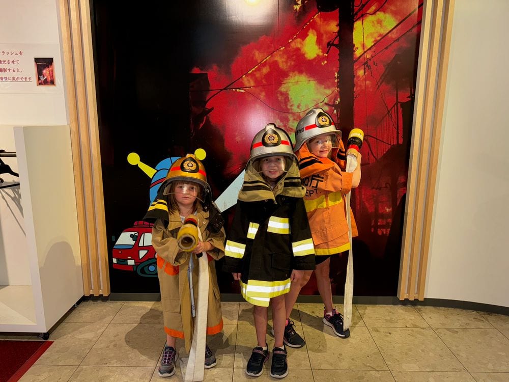 Three young children wearing yellow and orange firefighter costumes and helmets stand in front of a Tokyo Fire Department exhibit display.