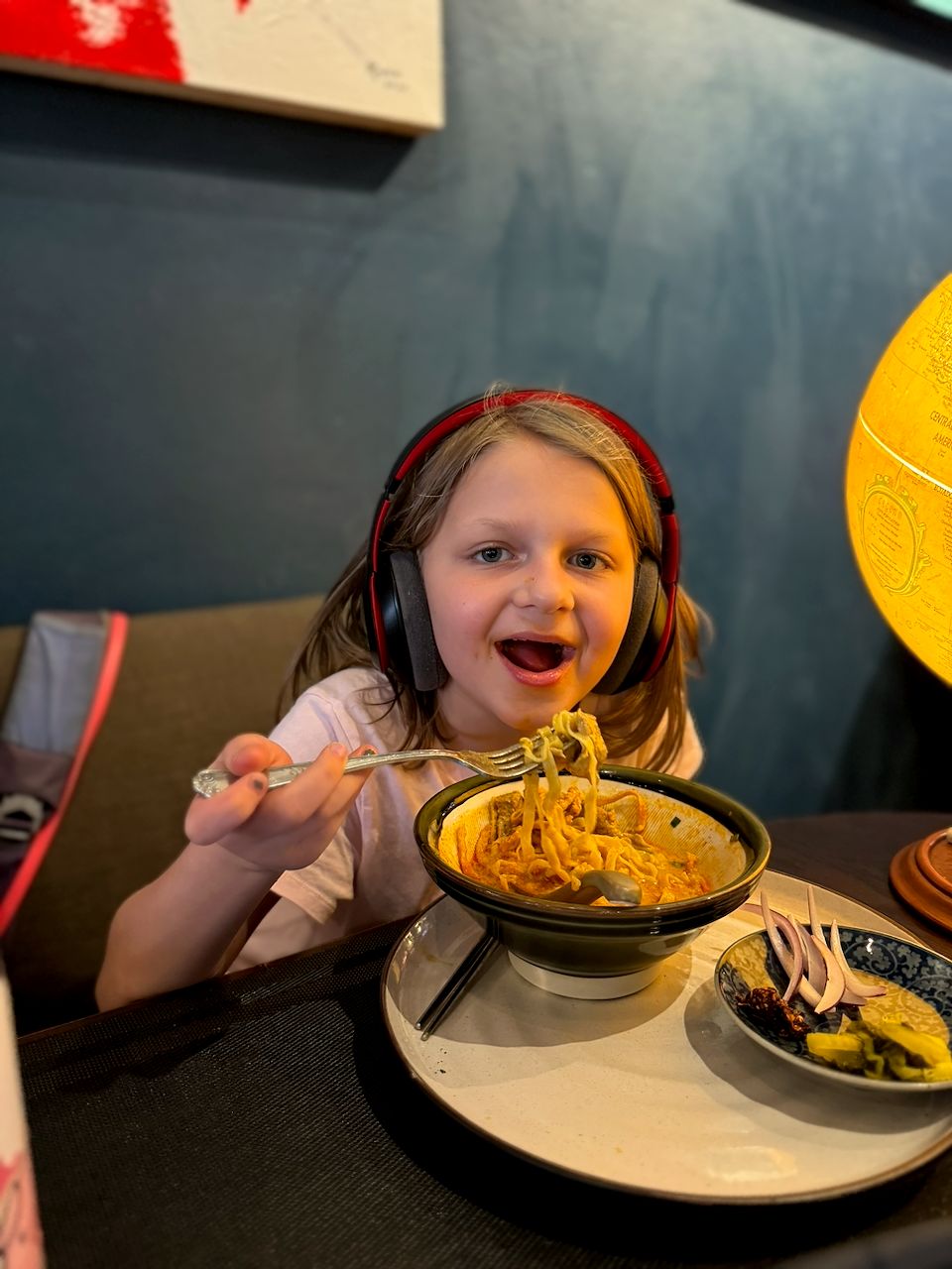 Cora sits at a restaurant table in Chiang Mai, wearing red headphones and grinning with her mouth open as she lifts a forkful of khao soi noodles from a bowl, with a small side dish of condiments next to it and a globe lamp visible in the background.