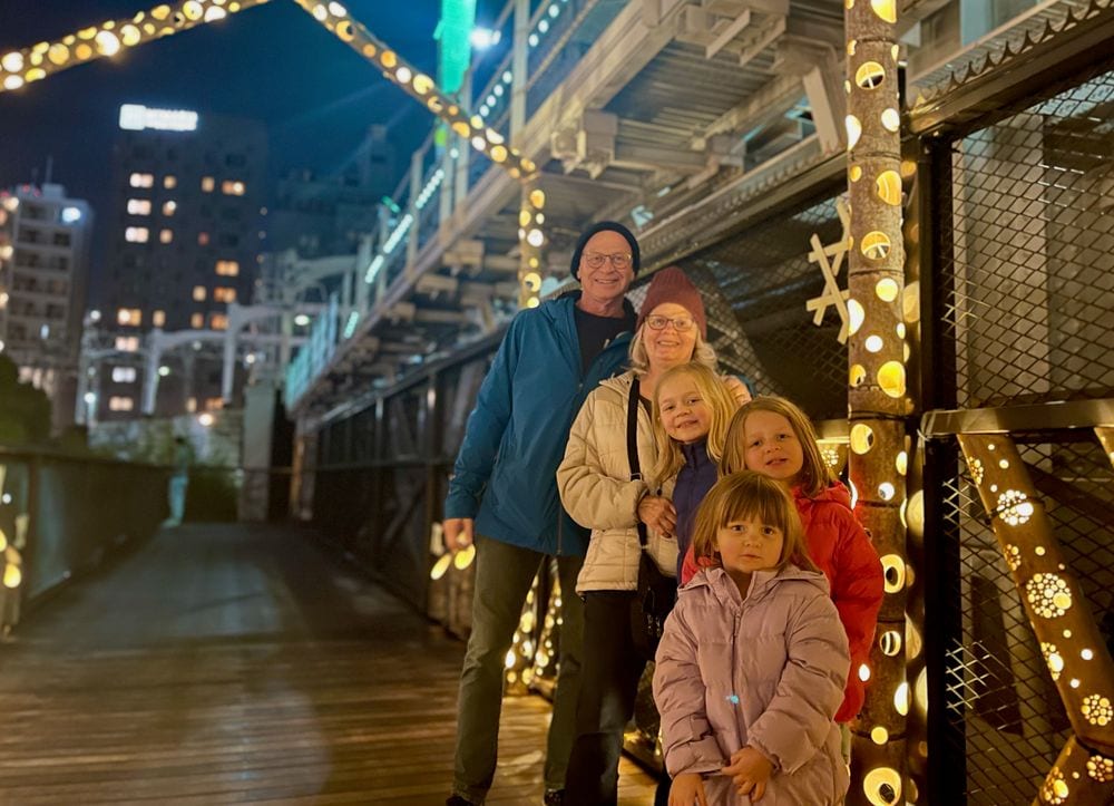 Two grandparents and three young girls pose together on a wooden pedestrian bridge decorated with warm glowing lights at night, with a city skyline visible in the background.