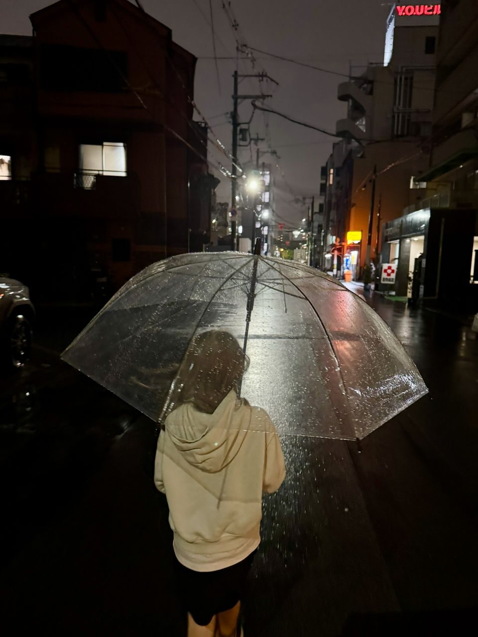 A person seen from behind holding a clear dome umbrella on a rain-slicked Osaka side street at night, with glowing signs visible in the distance.