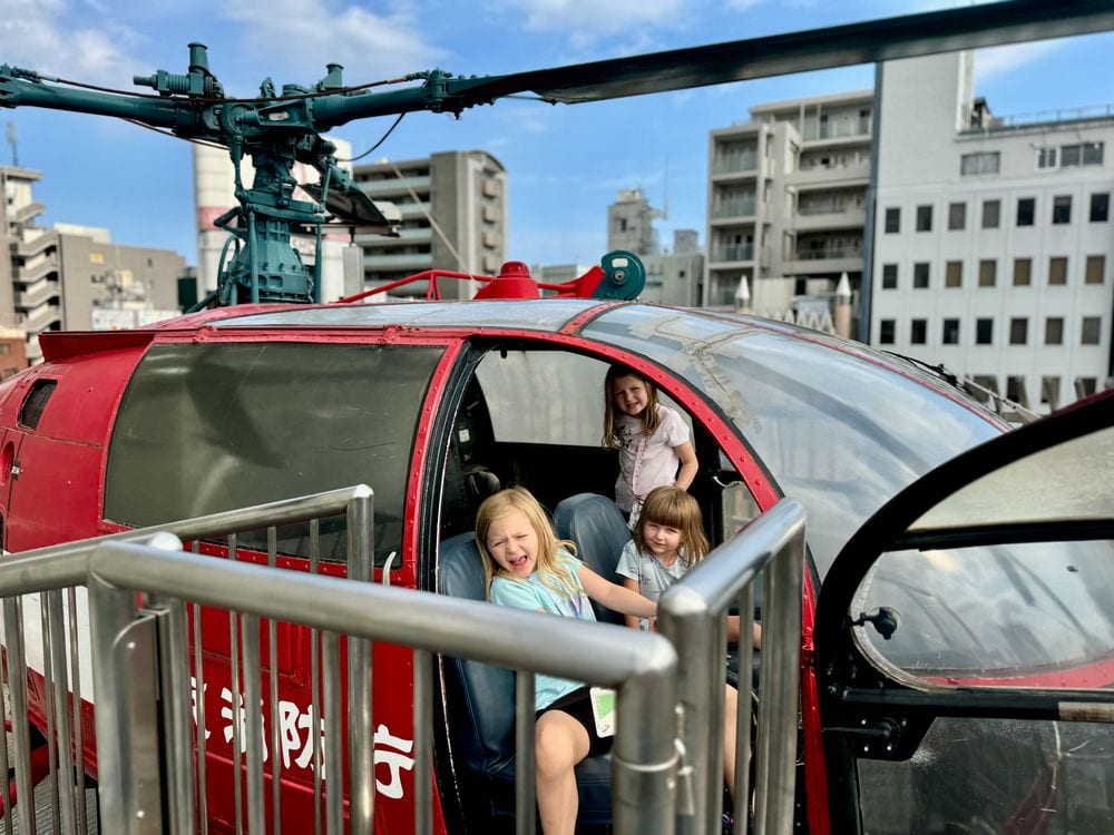 Two young girls sit inside the cockpit of a red fire department helicopter on the rooftop of the Tokyo Fire Museum.