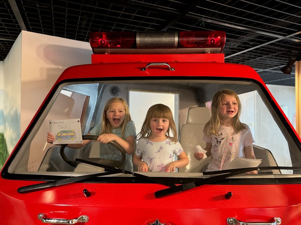Three young girls sit inside the cab of a red fire truck exhibit, all smiling and making excited expressions.