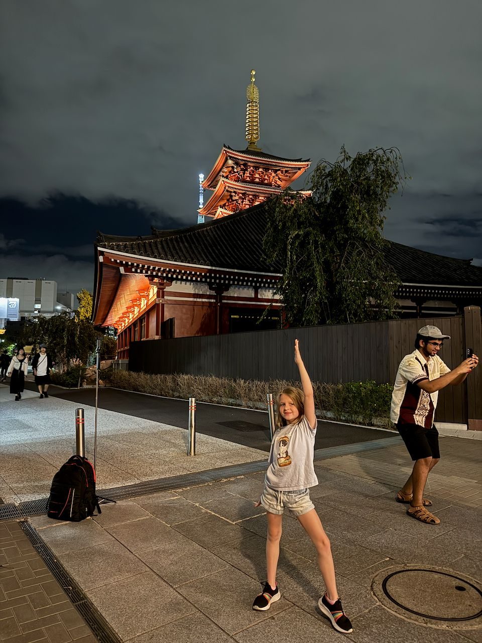 A young girl poses with one arm raised in front of the illuminated pagoda and main hall of Senso-ji temple at night in Asakusa, with the Tokyo Skytree visible in the background.