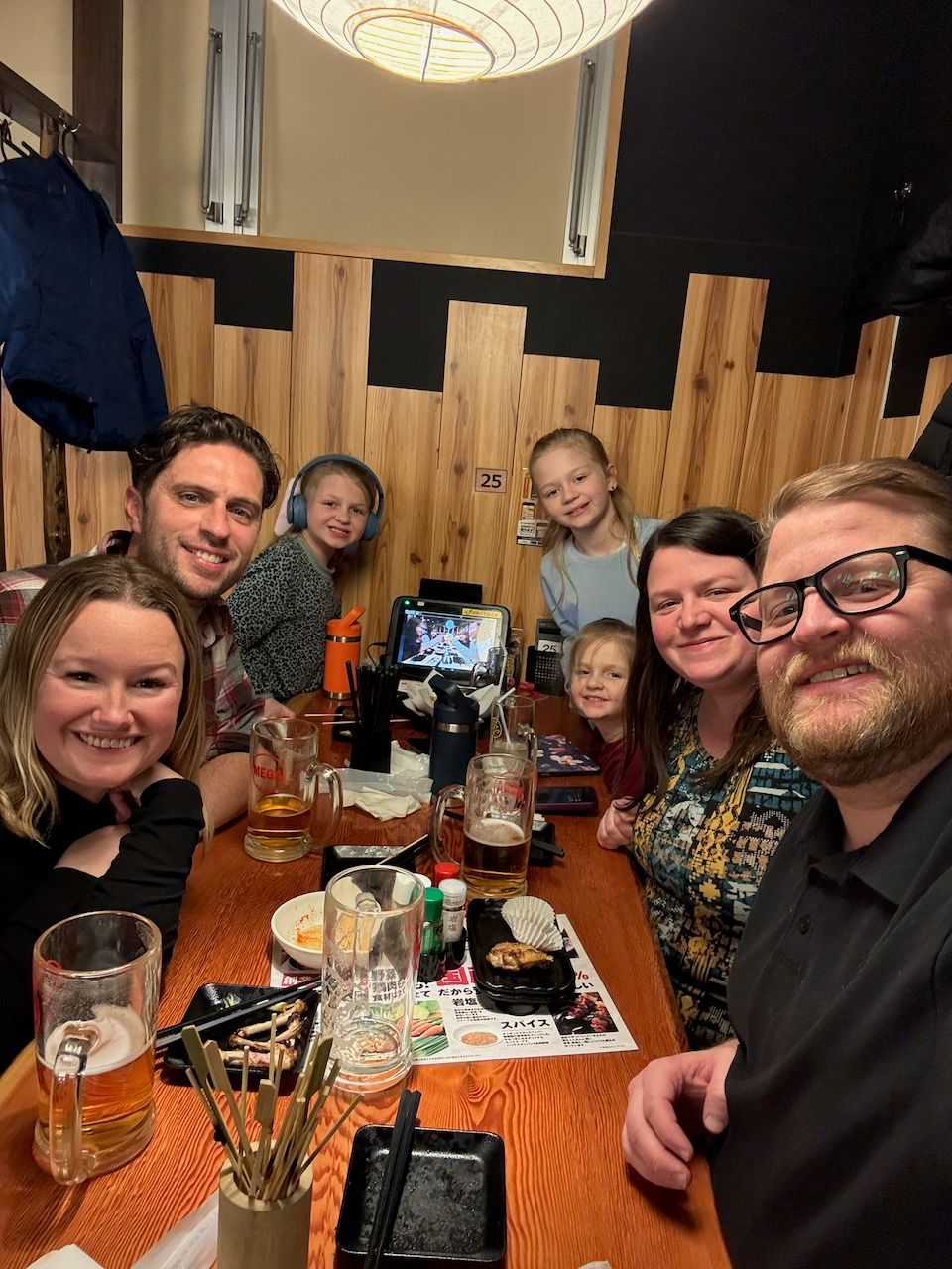 A group selfie of two families crammed into a wooden booth at a Japanese yakitori restaurant, with beer glasses, skewer trays, and chopstick holders covering the table and three kids visible in the background.