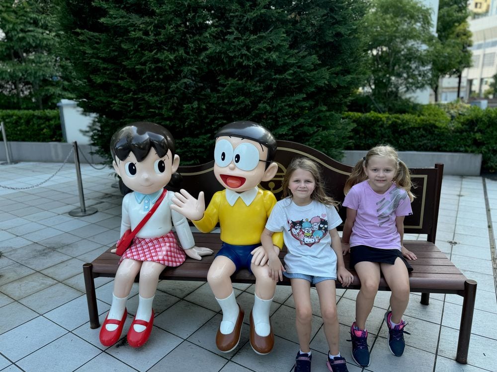Three young girls sit on a bench next to statues of Nobita and Shizuka from Doraemon, greenery visible behind them.