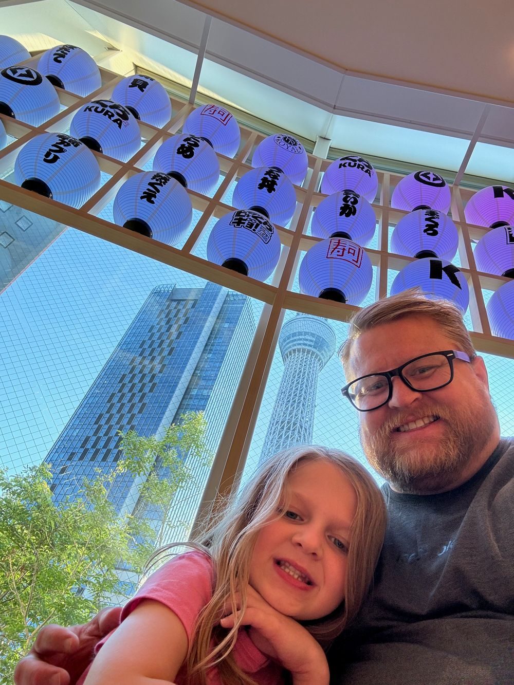 Purple lanterns bearing the Kura Sushi logo hang above a glass walkway with Tokyo Skytree visible through the window behind a father and daughter.