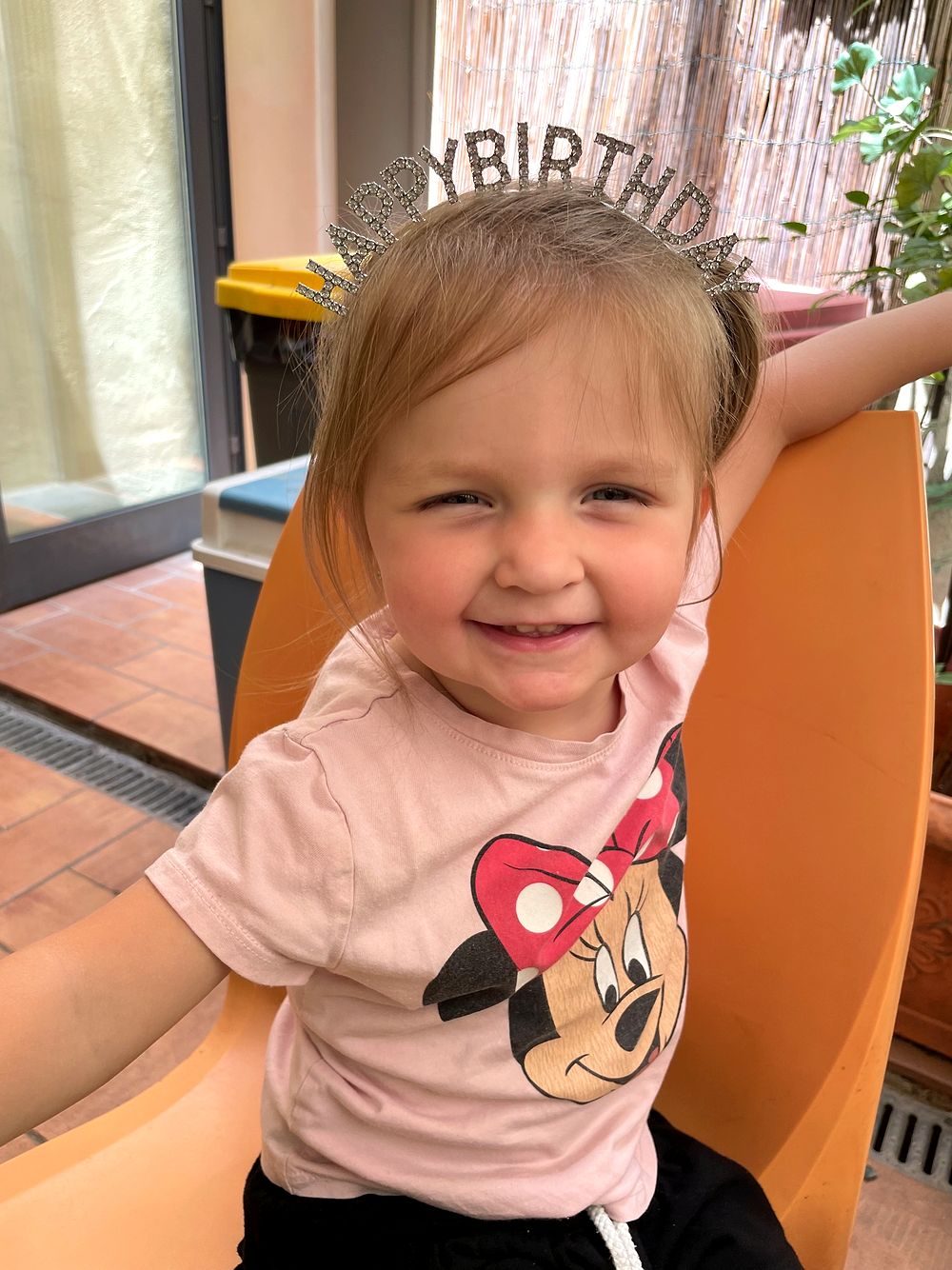A toddler girl sits in an orange chair wearing a sparkly Happy Birthday tiara and a pink Minnie Mouse t-shirt, grinning at the camera in a Tuscan apartment