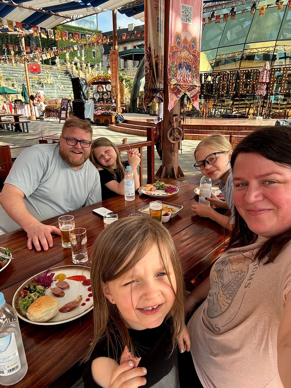 A family of five seated at a wooden picnic table at an outdoor beer festival venue with a carnival stage and bunting in the background, plates of sausage and salad in front of them, taking a selfie together.