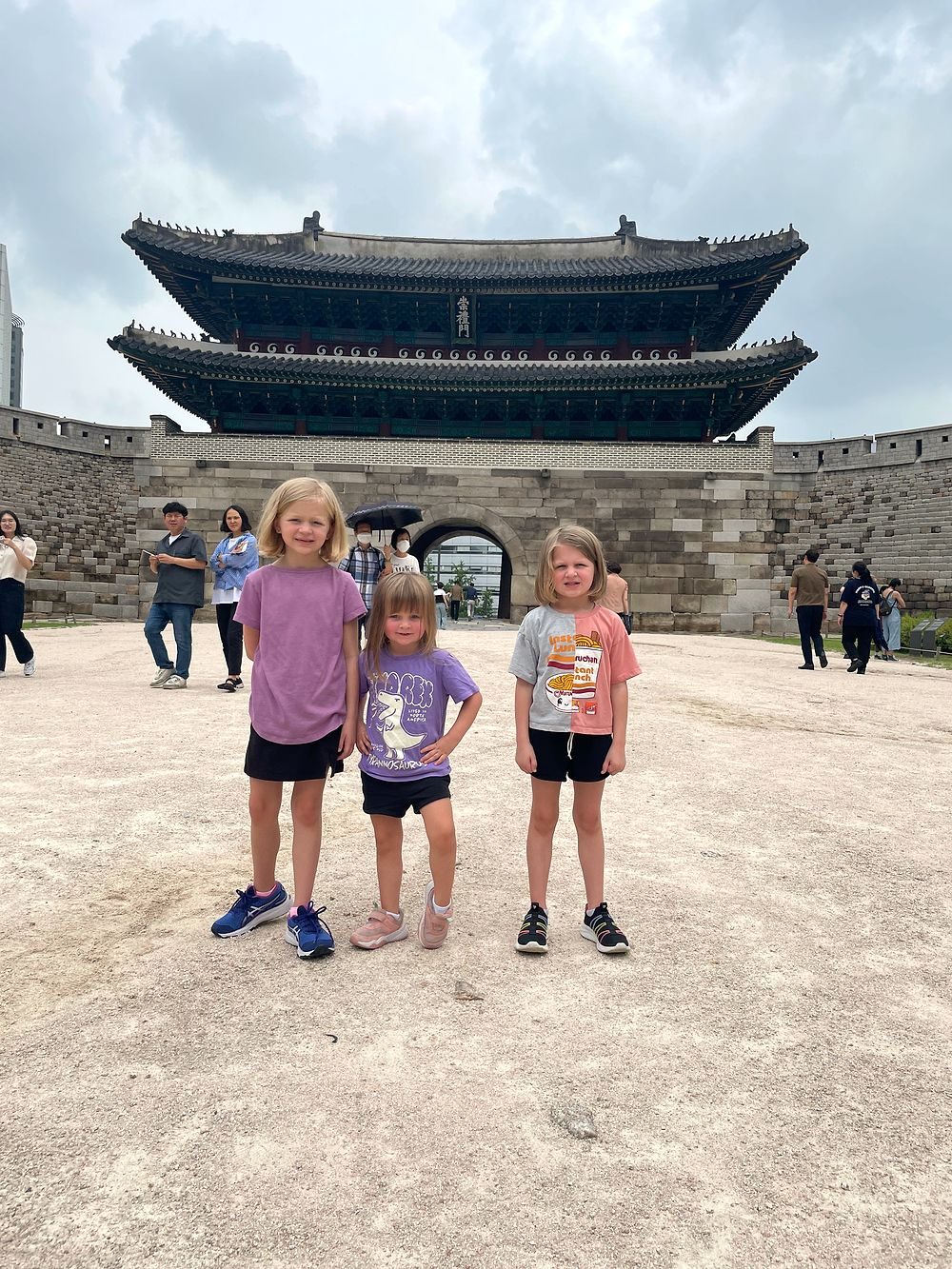 Three young girls standing in front of Namdaemun Gate in Seoul under a cloudy sky